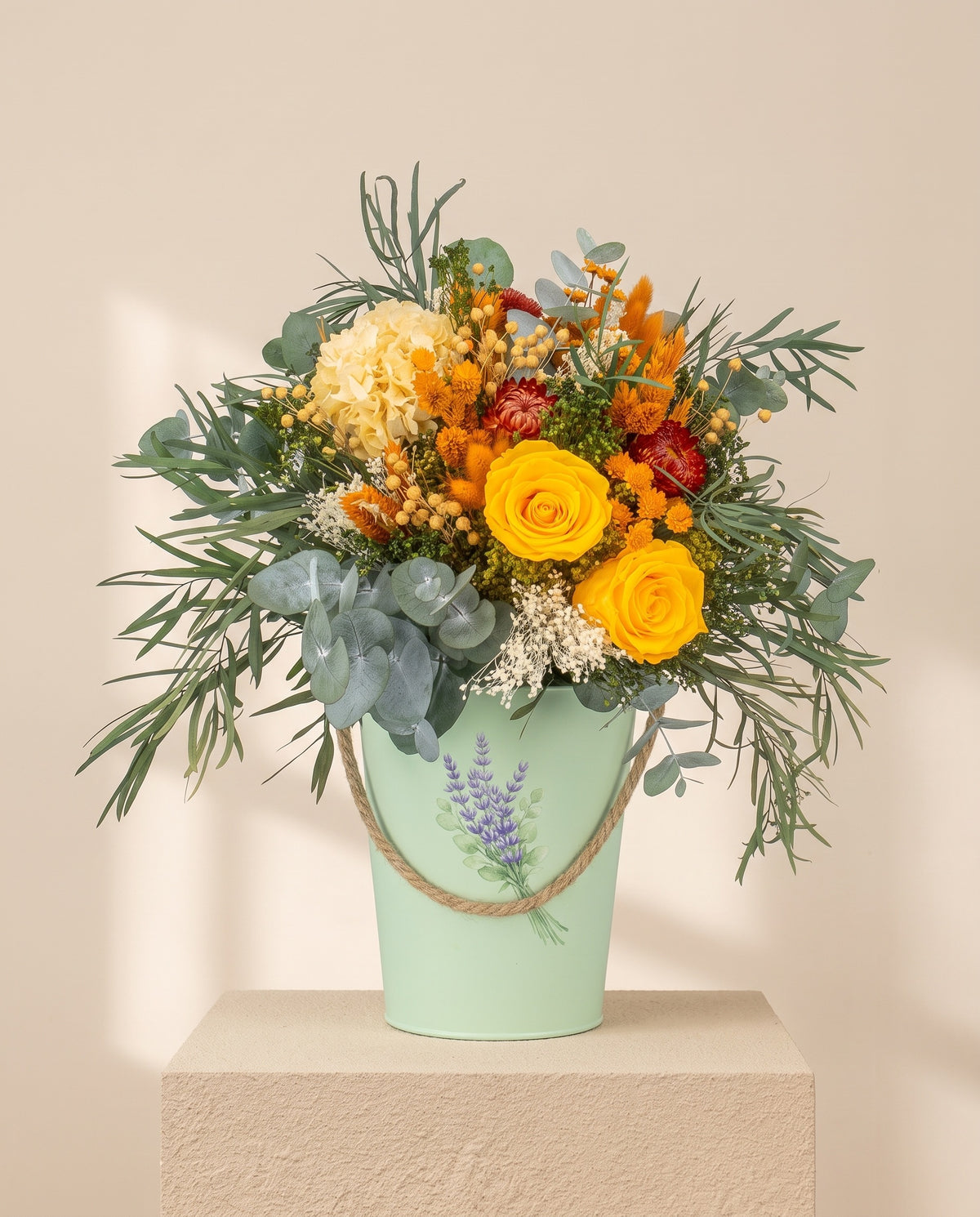 Centro de flores secas y preservadas en amarillo y naranja, con su recipiente, sobre fondo crema.