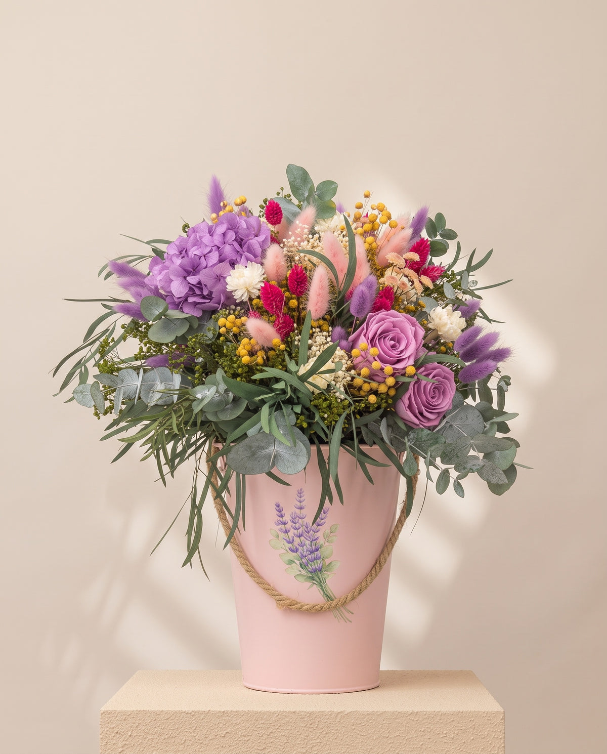 Centro de flores secas e preservadas em púrpura e cor-de-rosa, com o seu recipiente, sobre fundo creme.