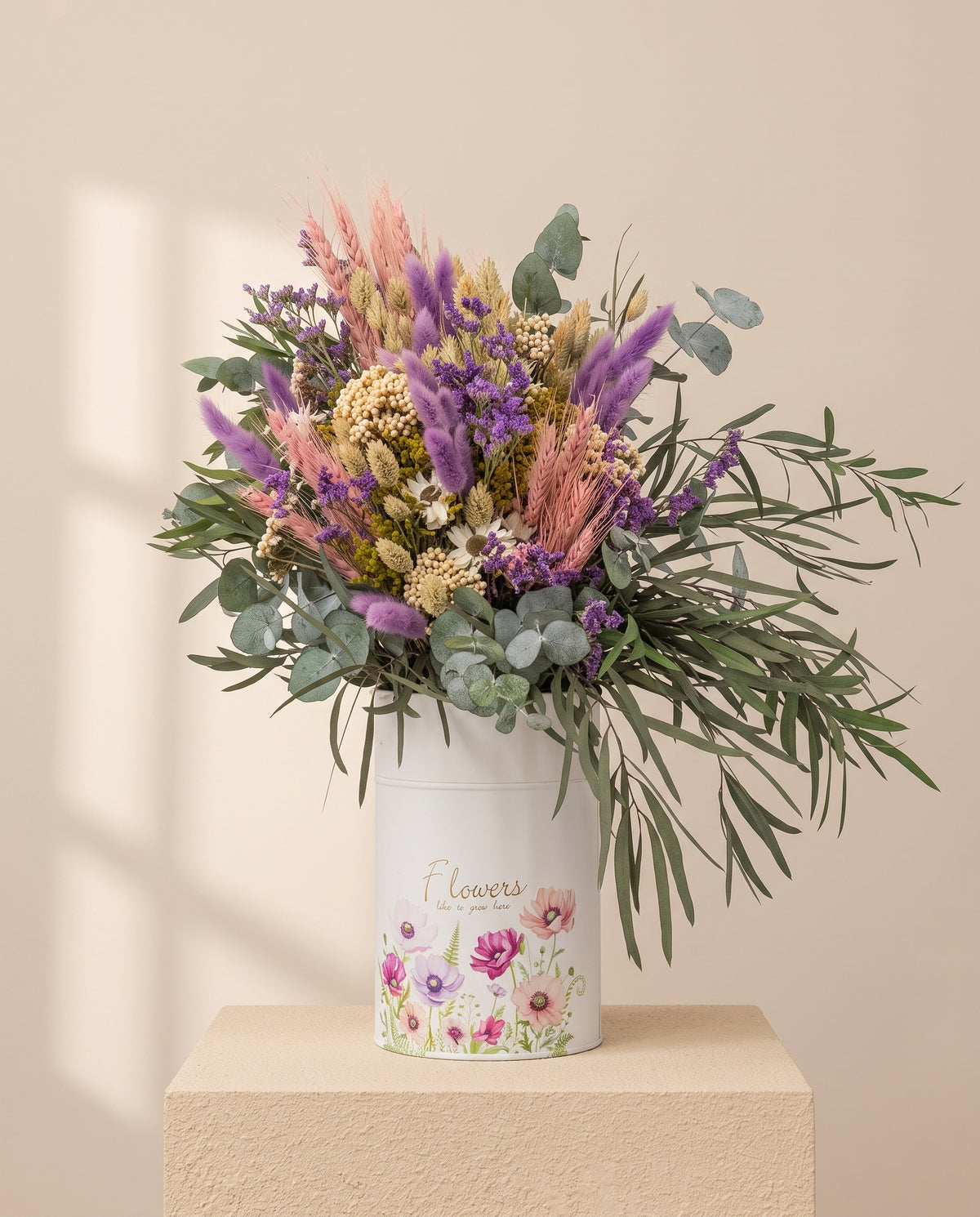 Centro de flores secas e preservadas em púrpura e cor-de-rosa, com o seu recipiente, sobre fundo creme.