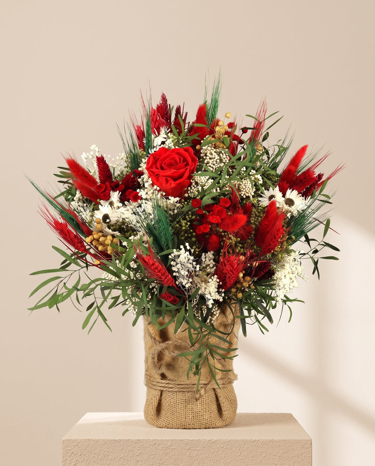 Centro de flores secas y preservadas en rojo, con su recipiente, sobre fondo crema.