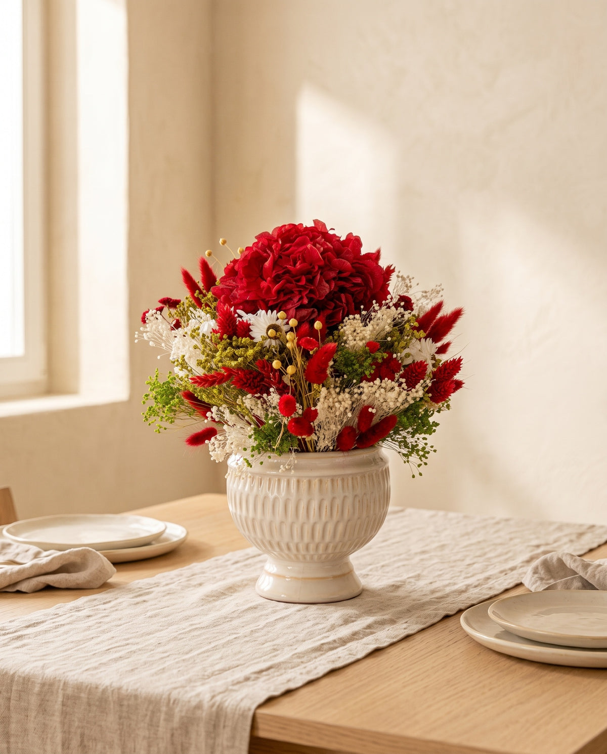 Centro de flores secas e preservadas em vermelho e branco numa mesa de sala de jantar.