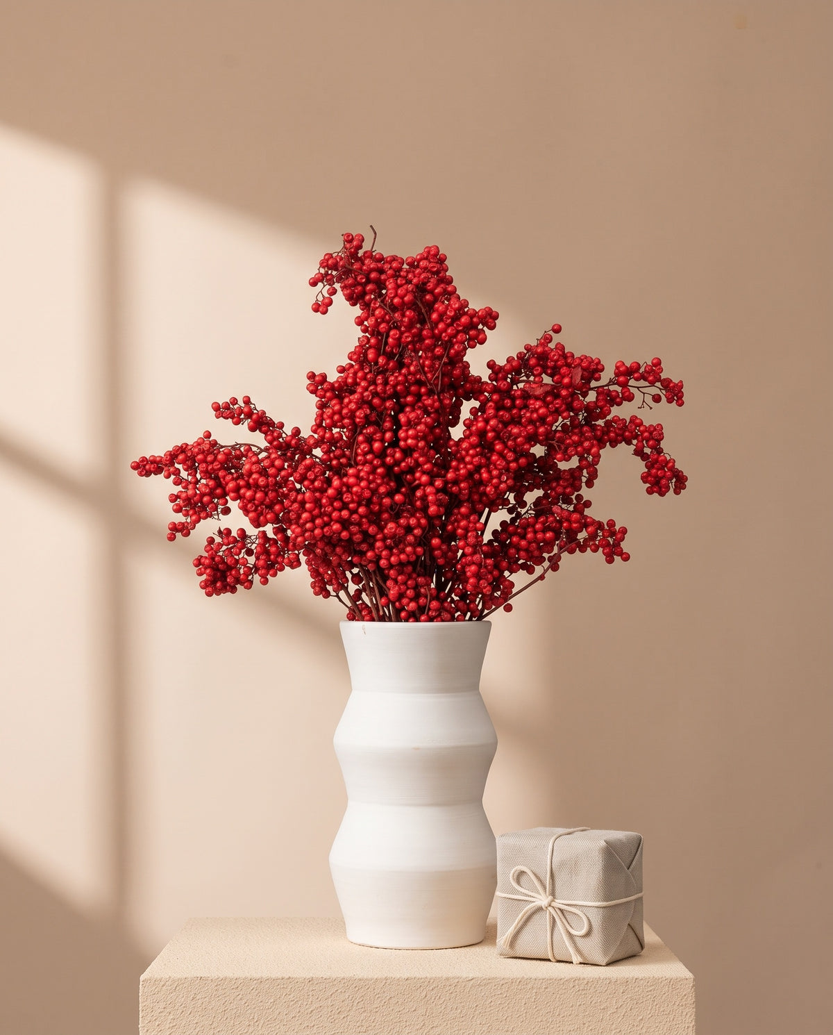 Ramas de falsa pimienta con bayas rojas en jarrón de cerámica blanca, con caja regalo de lino y pared camel. Jarrón y caja no incluidos.