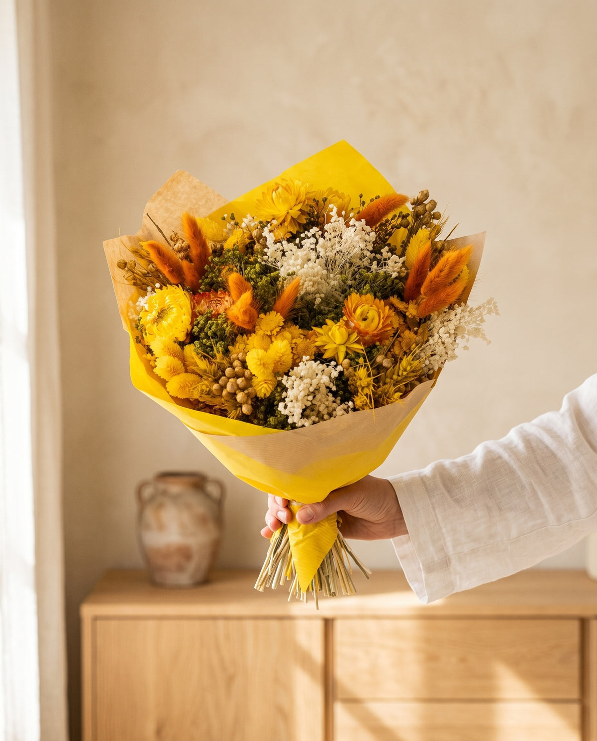 Mão a segurar o ramo de flores secas em amarelo com o seu papel, tal como é entregue.