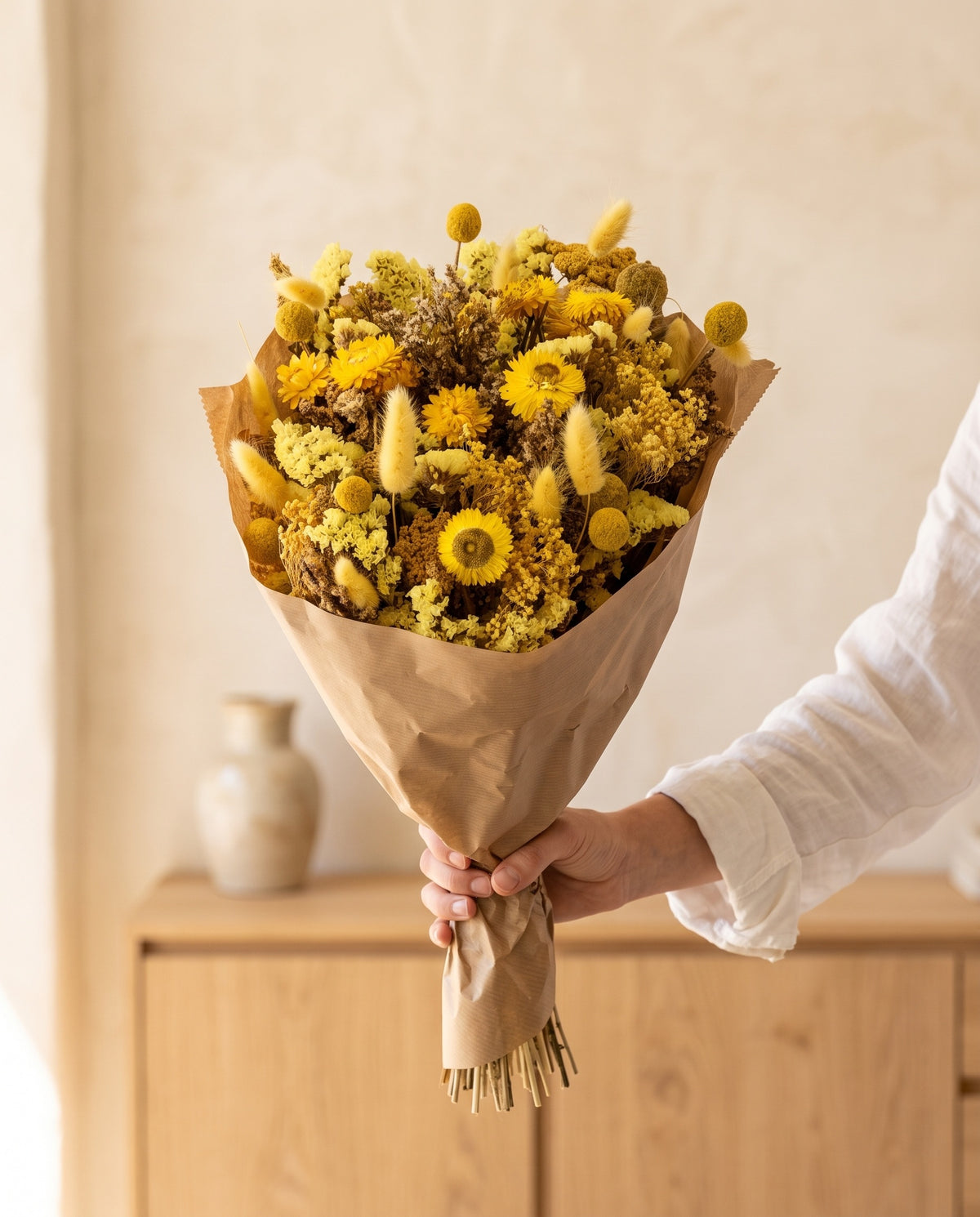 Mão a segurar o ramo grande de flores secas em amarelo com o seu papel, tal como é entregue.
