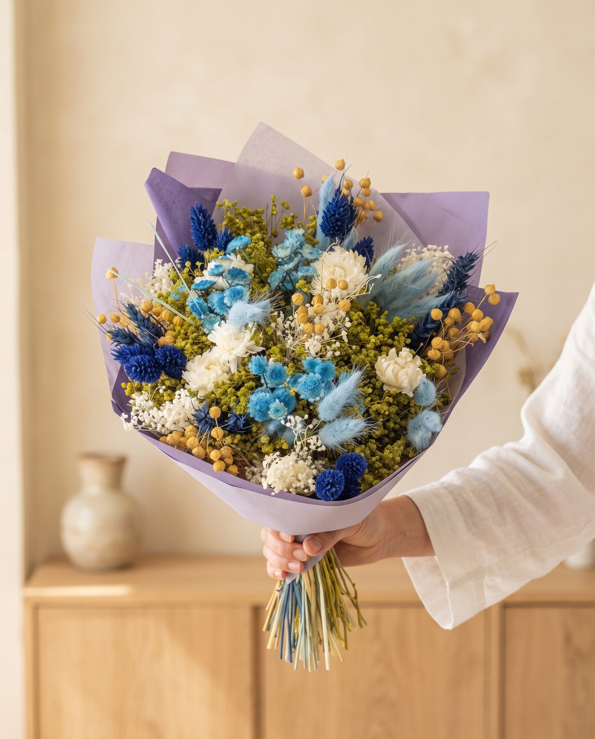 Mão a segurar o ramo de flores secas em azul com o seu papel, tal como é entregue.