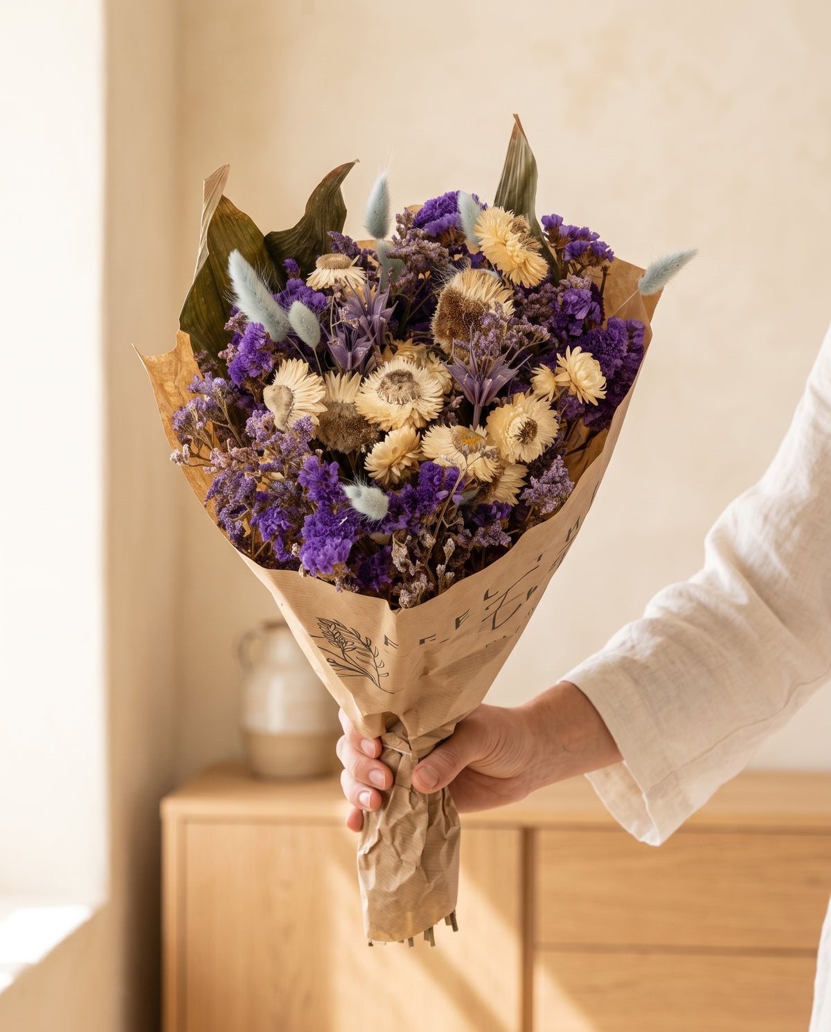 Mão a segurar o ramo de flores secas em azul com o seu papel, tal como é entregue.