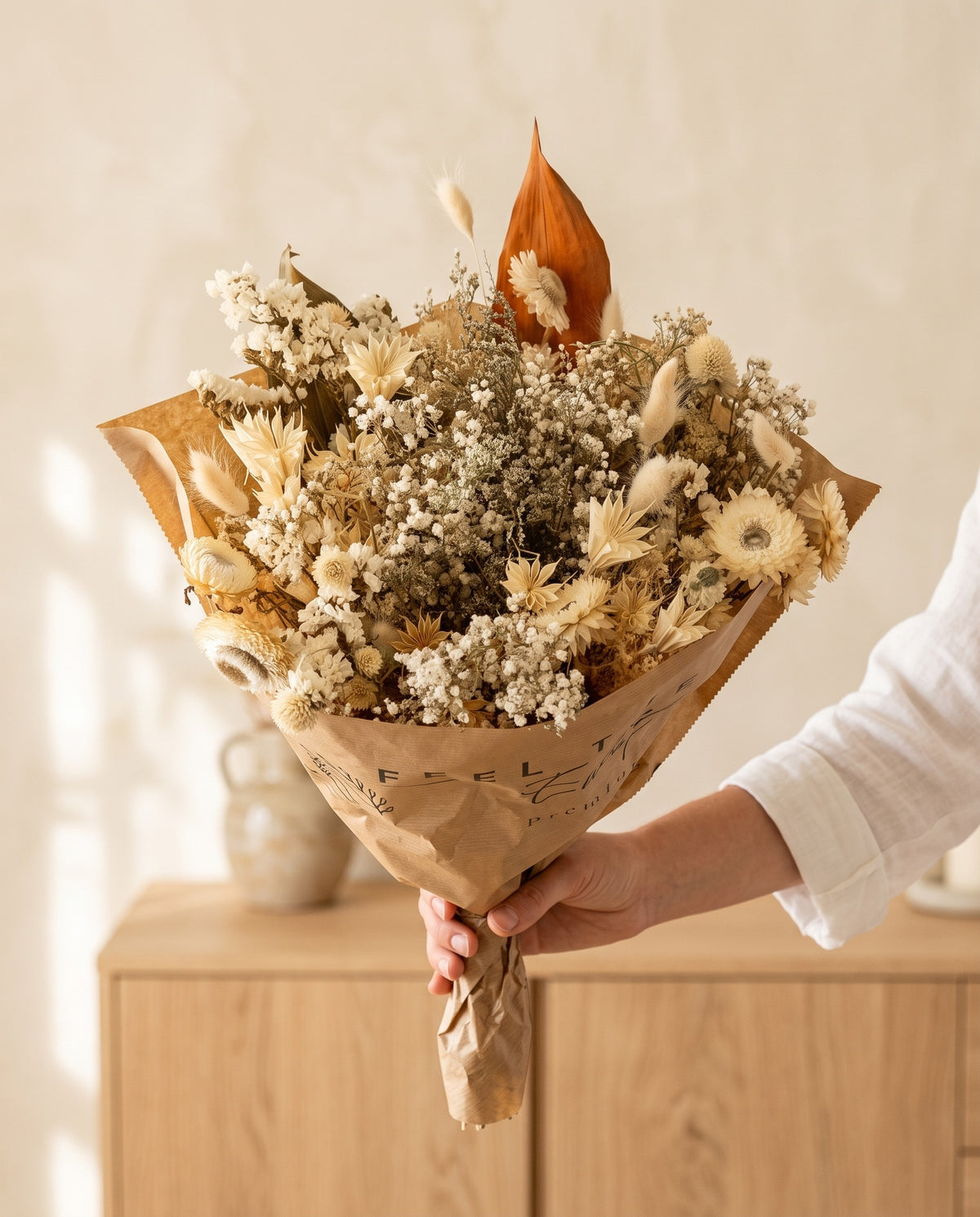 Mão a segurar o ramo grande de flores secas em branco com o seu papel, tal como é entregue.