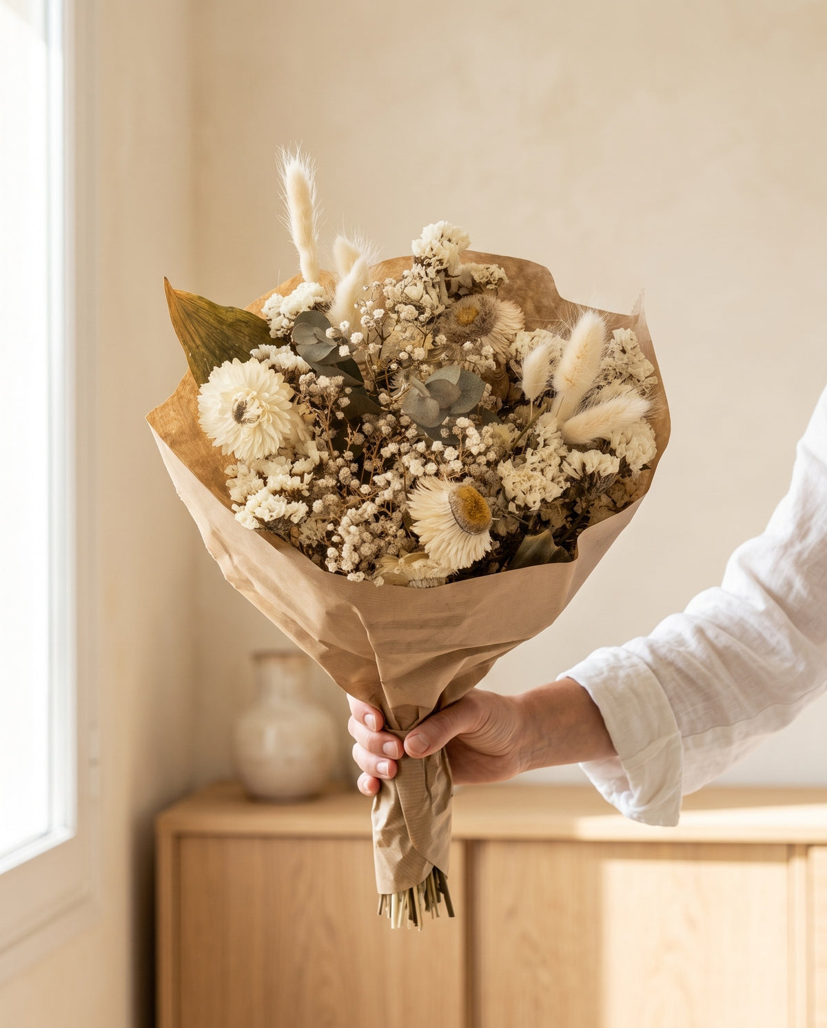 Mão a segurar o ramo de flores secas em branco com o seu papel, tal como é entregue.