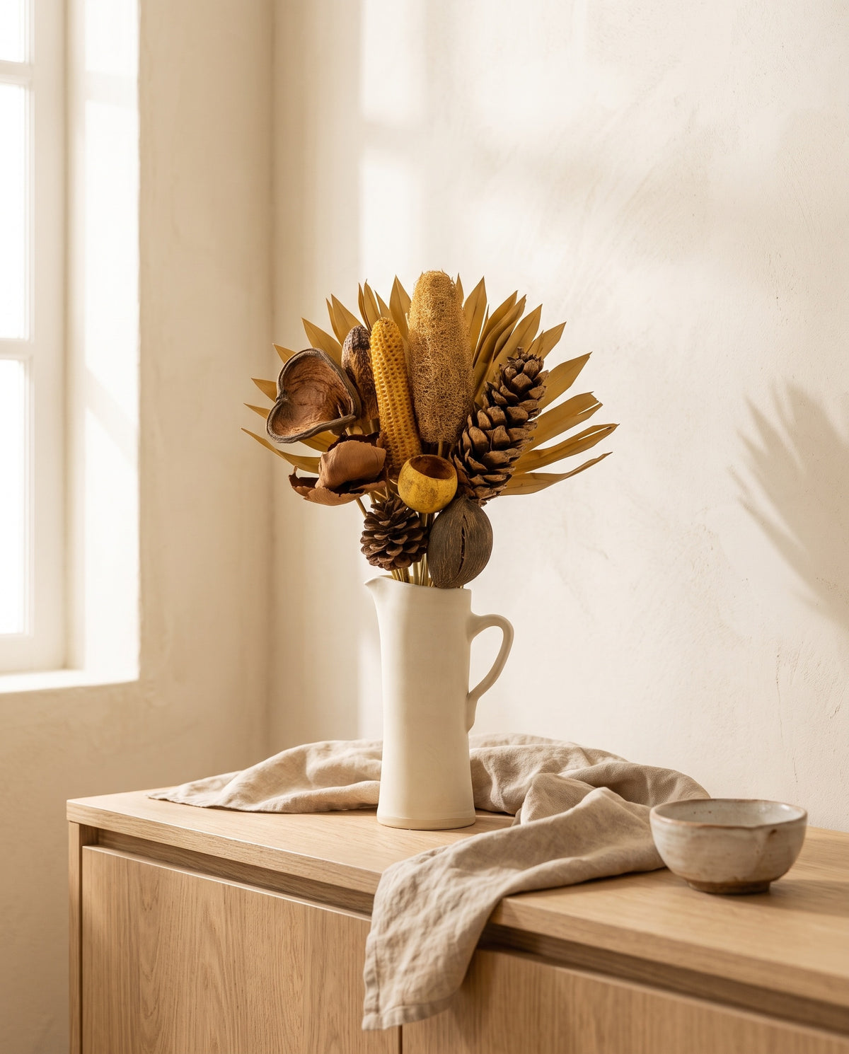 Ramo de flores secas em creme com pinhas numa jarra sobre um aparador, ambiente de casa. Jarra não incluída.