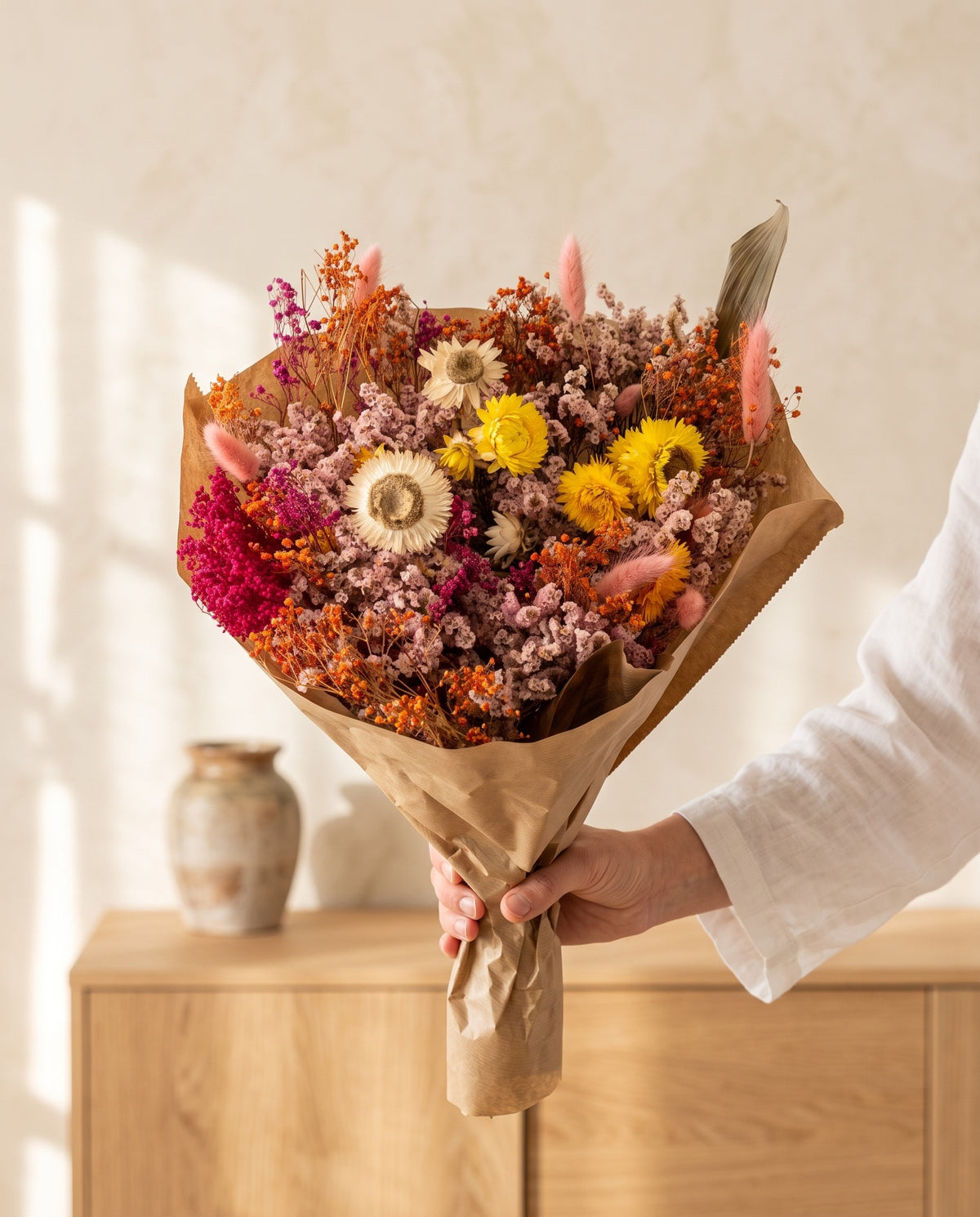 Mão a segurar o ramo grande de flores secas multicolor com o seu papel, tal como é entregue.