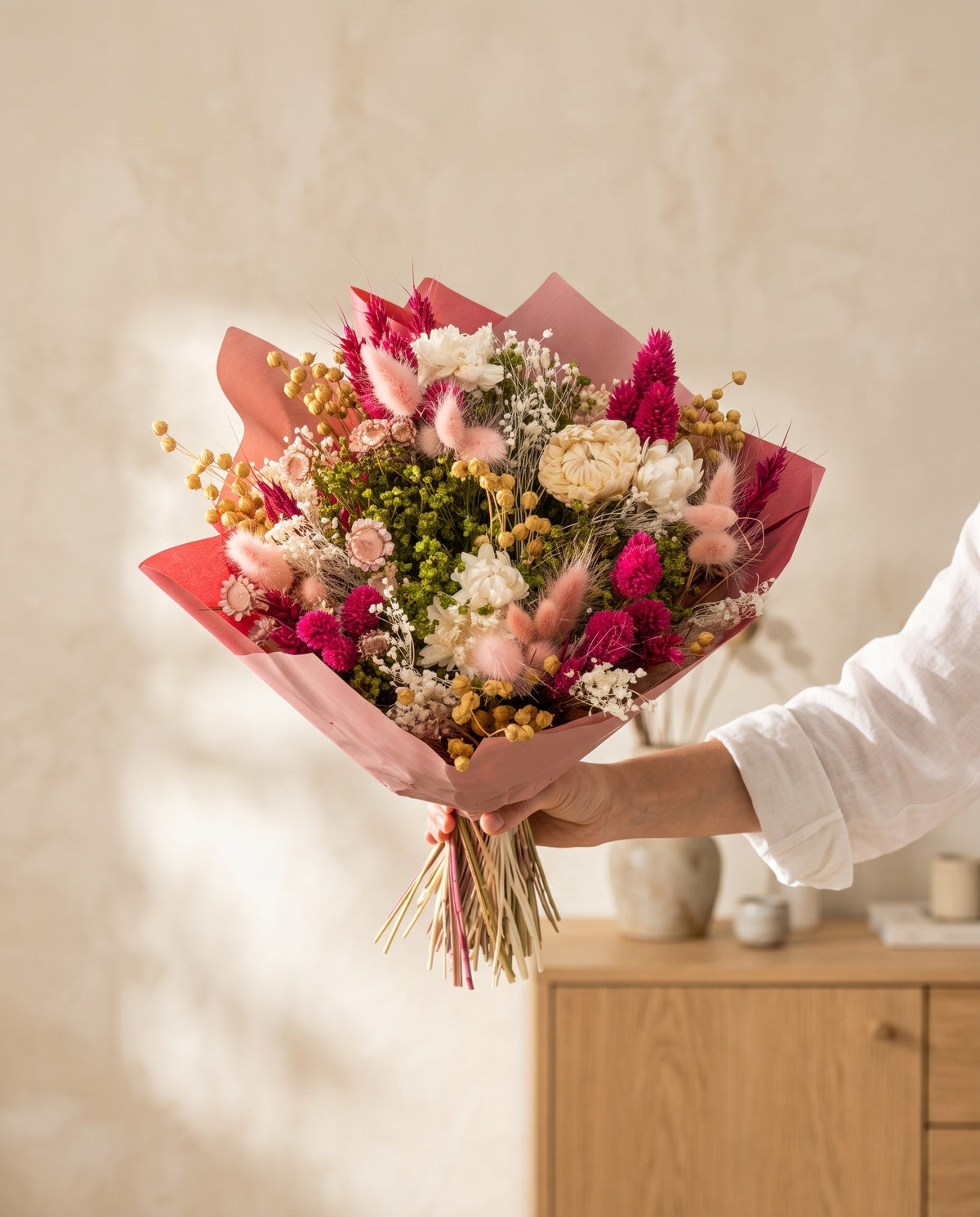 Mão a segurar o ramo de flores secas em fúcsia com phalaris com o seu papel, tal como é entregue.