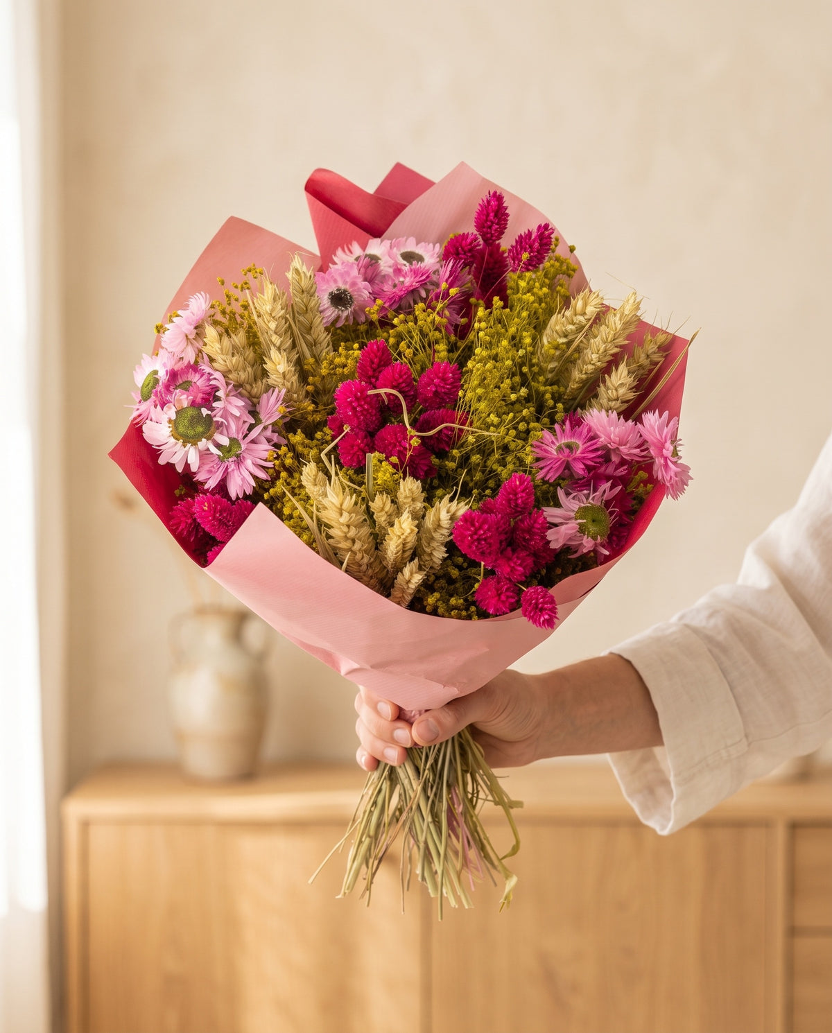Mão a segurar o ramo de flores secas em fúcsia com trigo com o seu papel, tal como é entregue.