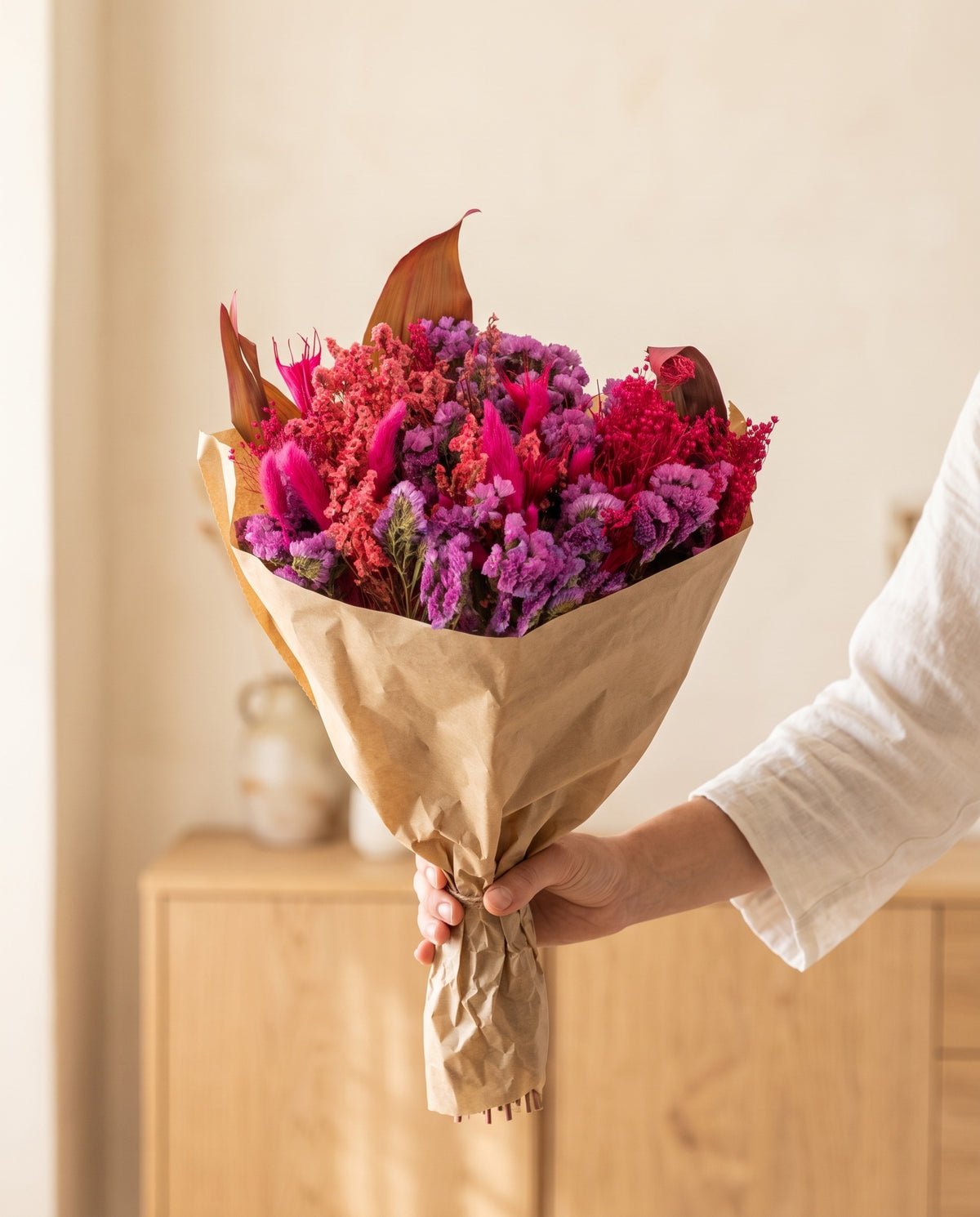 Mão a segurar o ramo de flores secas em fúcsia com o seu papel, tal como é entregue.