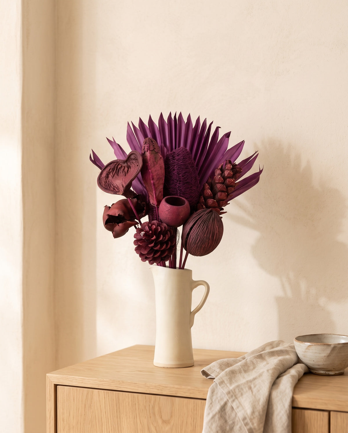 Ramo de flores secas en morado con piñas en una jarra sobre un aparador, ambiente de casa. Jarra no incluida.
