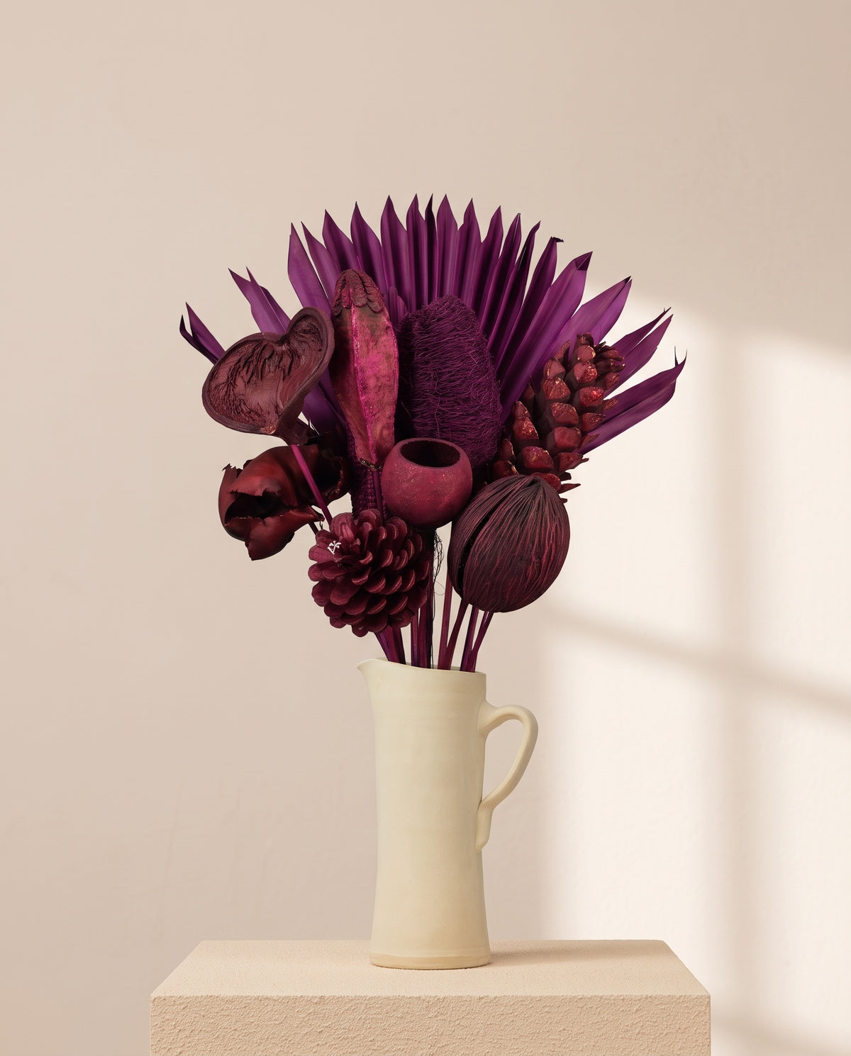 Ramo de flores secas en morado con piñas en una jarra de cerámica crema. Jarra no incluida.