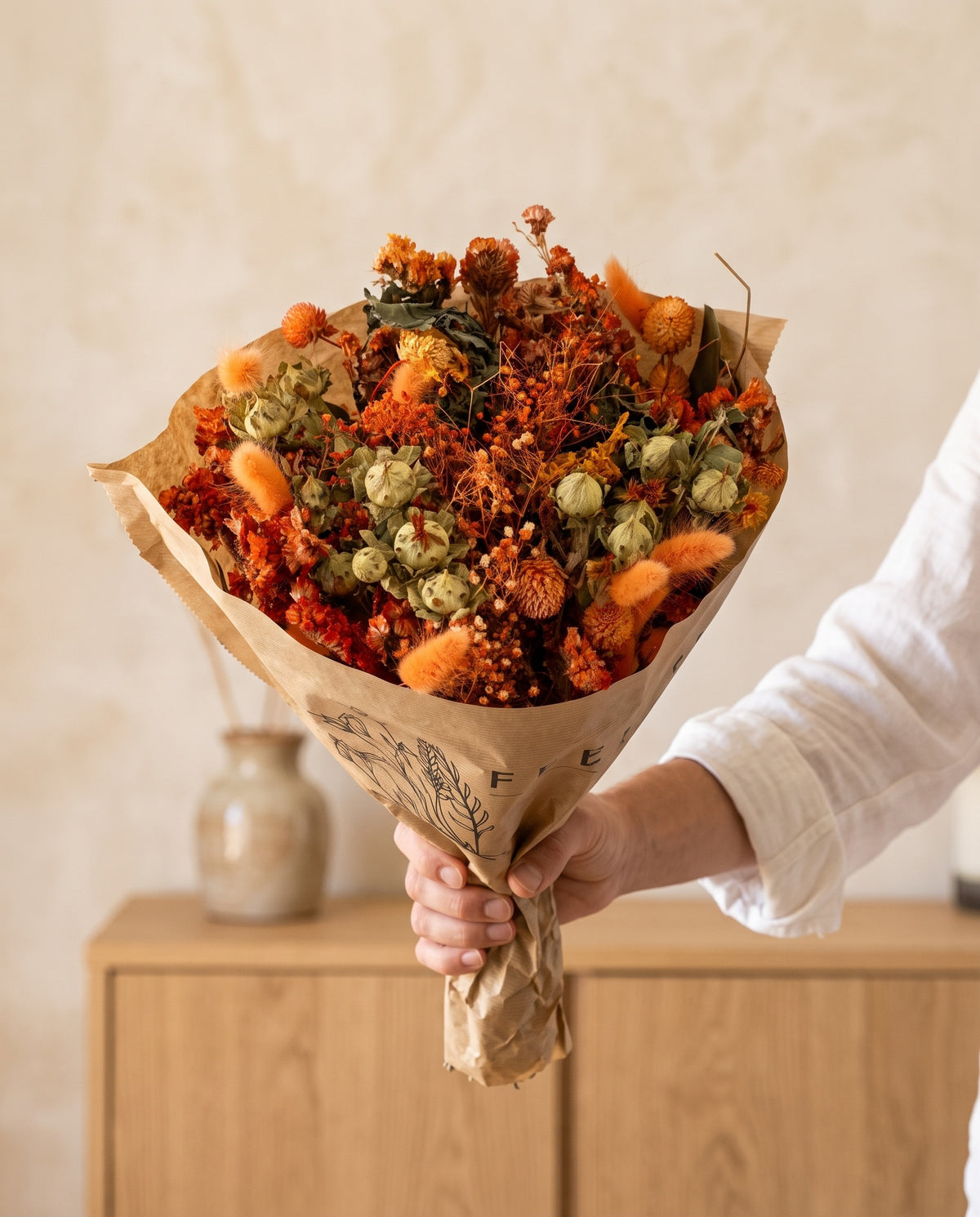 Mano sujetando el ramo grande de flores secas en naranja con su papel, como se entrega.