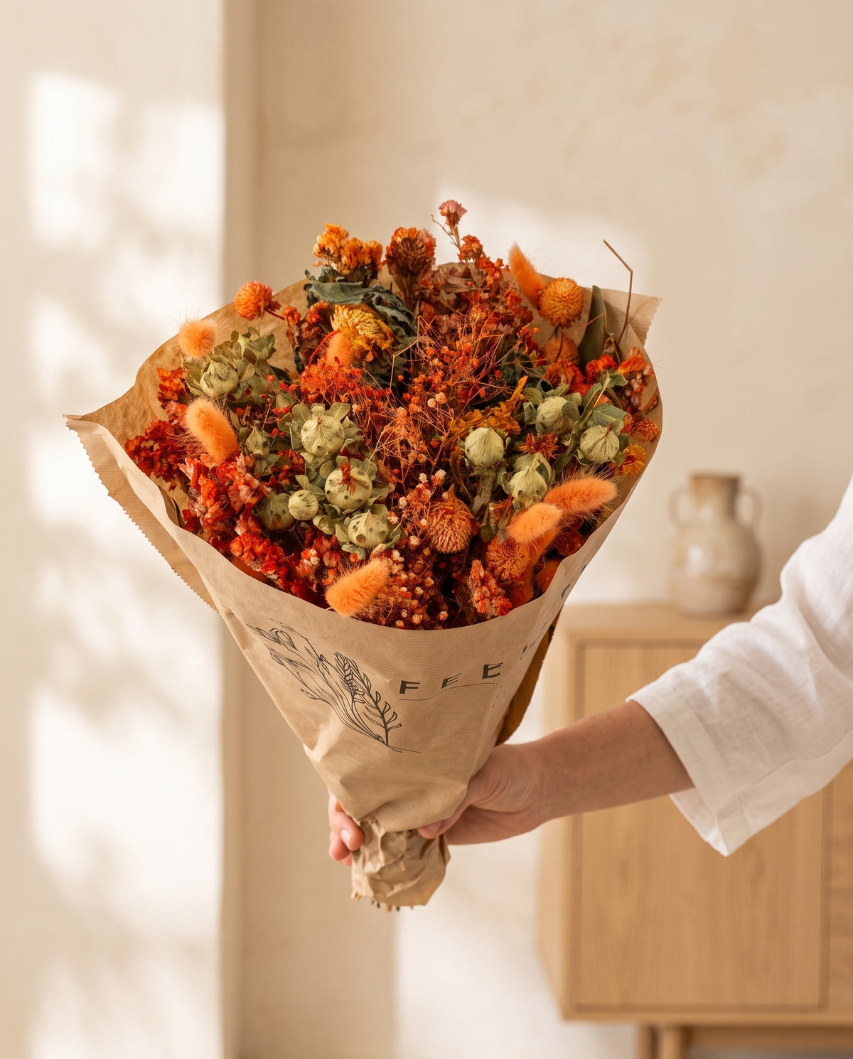 Mão a segurar o ramo de flores secas em laranja com o seu papel, tal como é entregue.