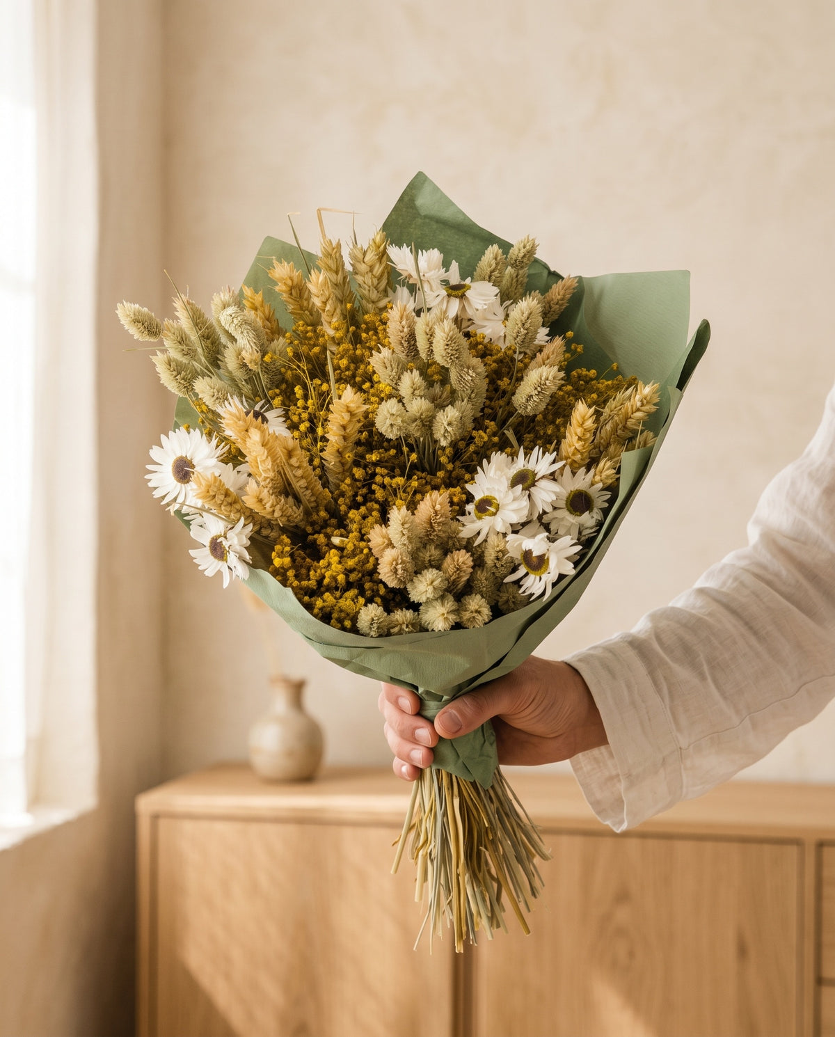 Mão a segurar o ramo de flores secas em tons naturais com phalaris com o seu papel, tal como é entregue.
