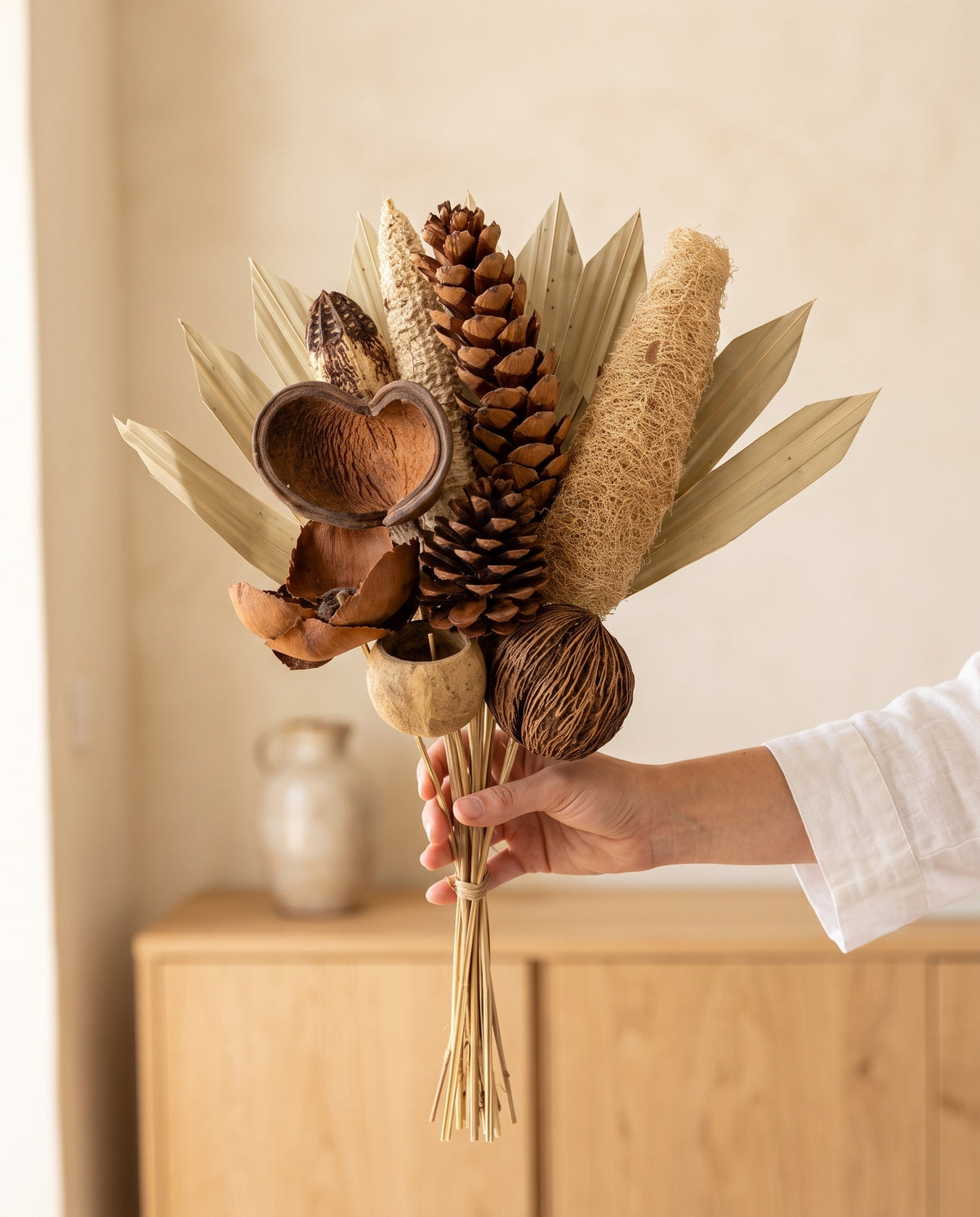 Mão a segurar o ramo de flores secas em tons naturais com pinhas, para ver o tamanho.