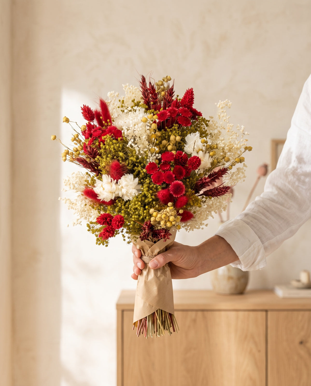 Mão a segurar o ramo de flores secas em vermelho com o seu papel, tal como é entregue.