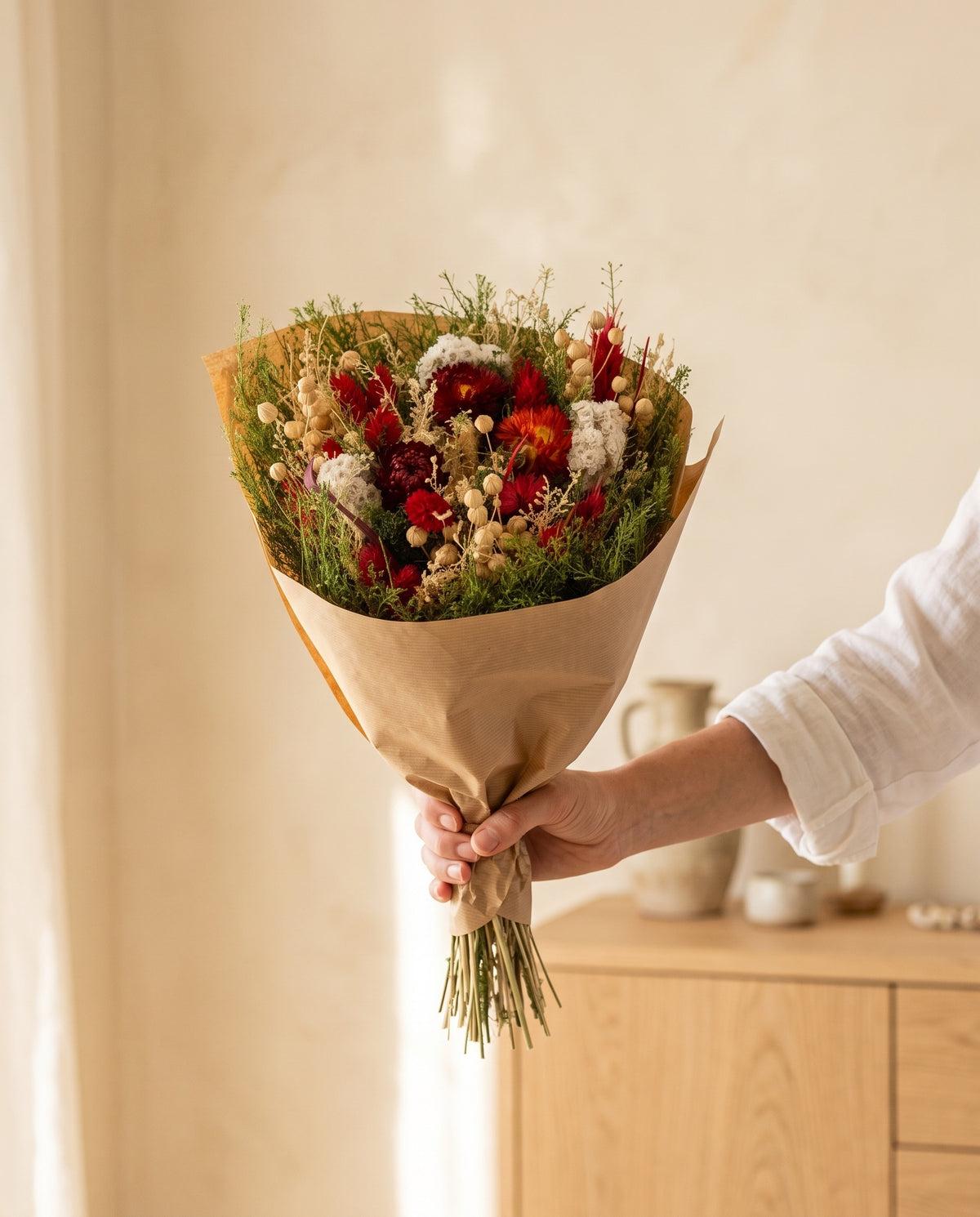 Mão a segurar o ramo de flores secas em vermelho com o seu papel, tal como é entregue.