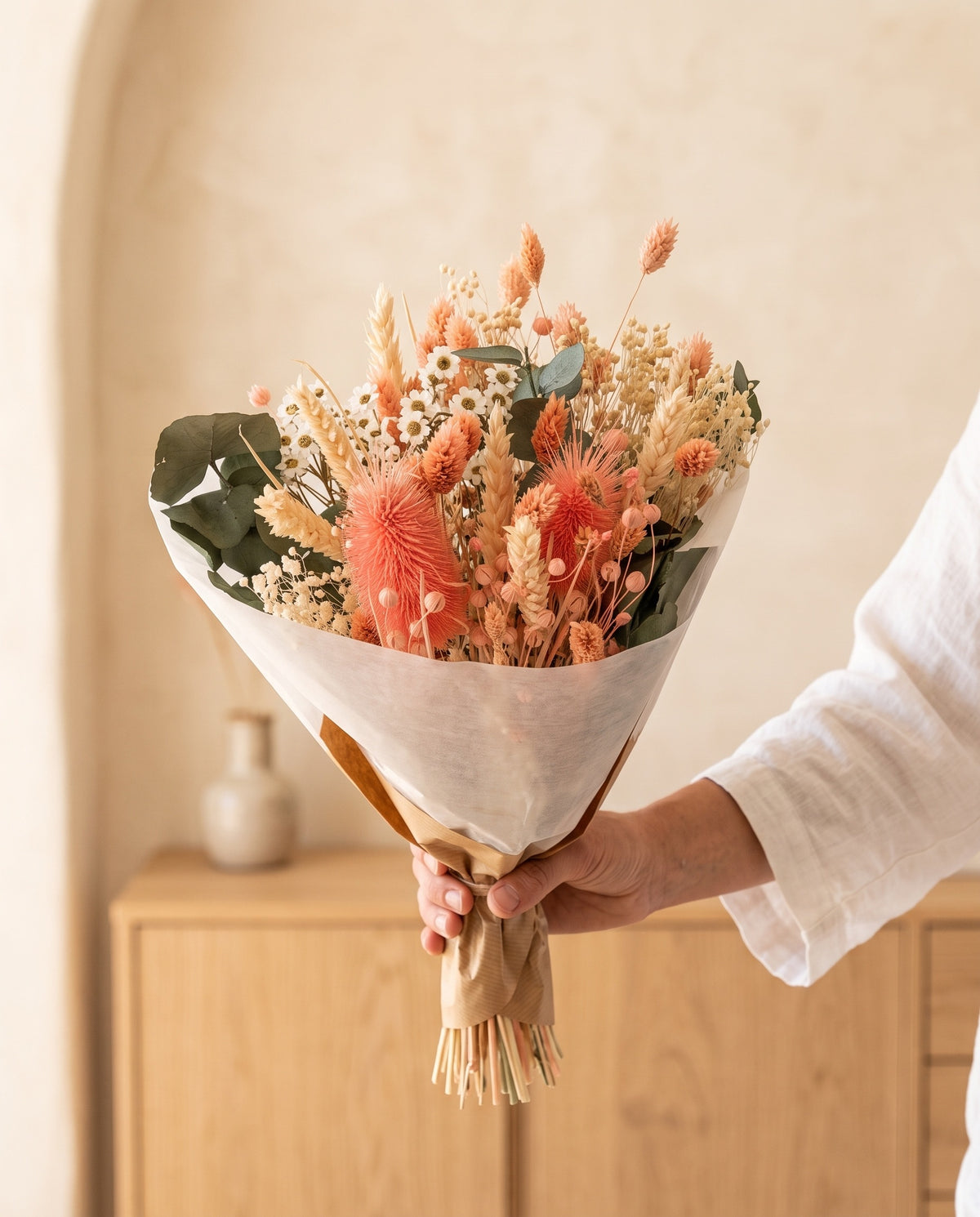 Mão a segurar o ramo de flores secas em rosa com o seu papel, tal como é entregue.