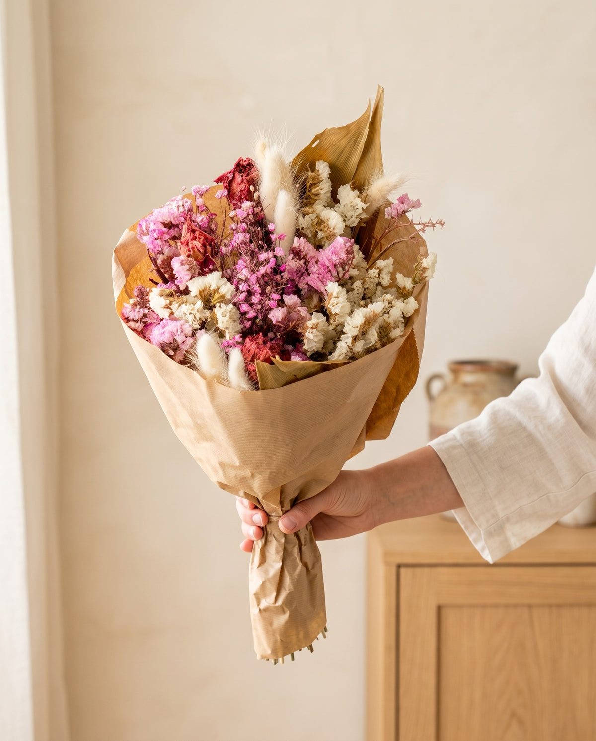 Mão a segurar o ramo de flores secas em rosa com o seu papel, tal como é entregue.