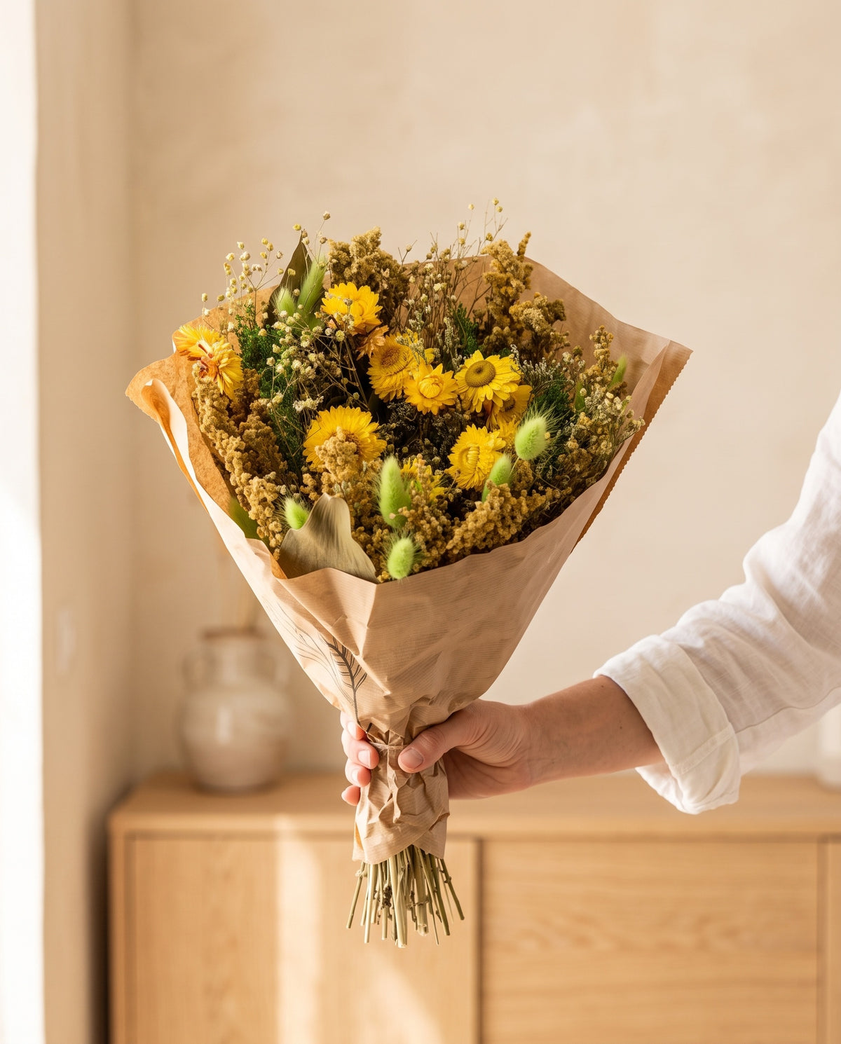 Mão a segurar o ramo grande de flores secas em verde e amarelo com o seu papel, tal como é entregue.