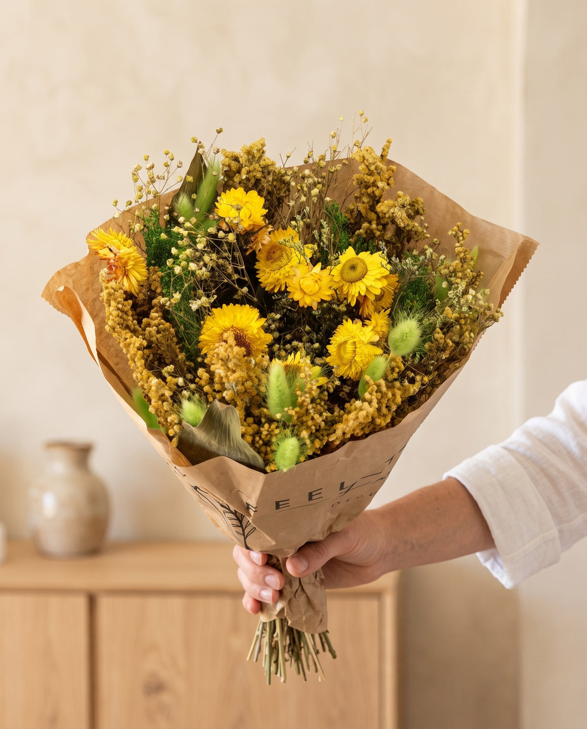 Mão a segurar o ramo de flores secas em verde e amarelo com o seu papel, tal como é entregue.