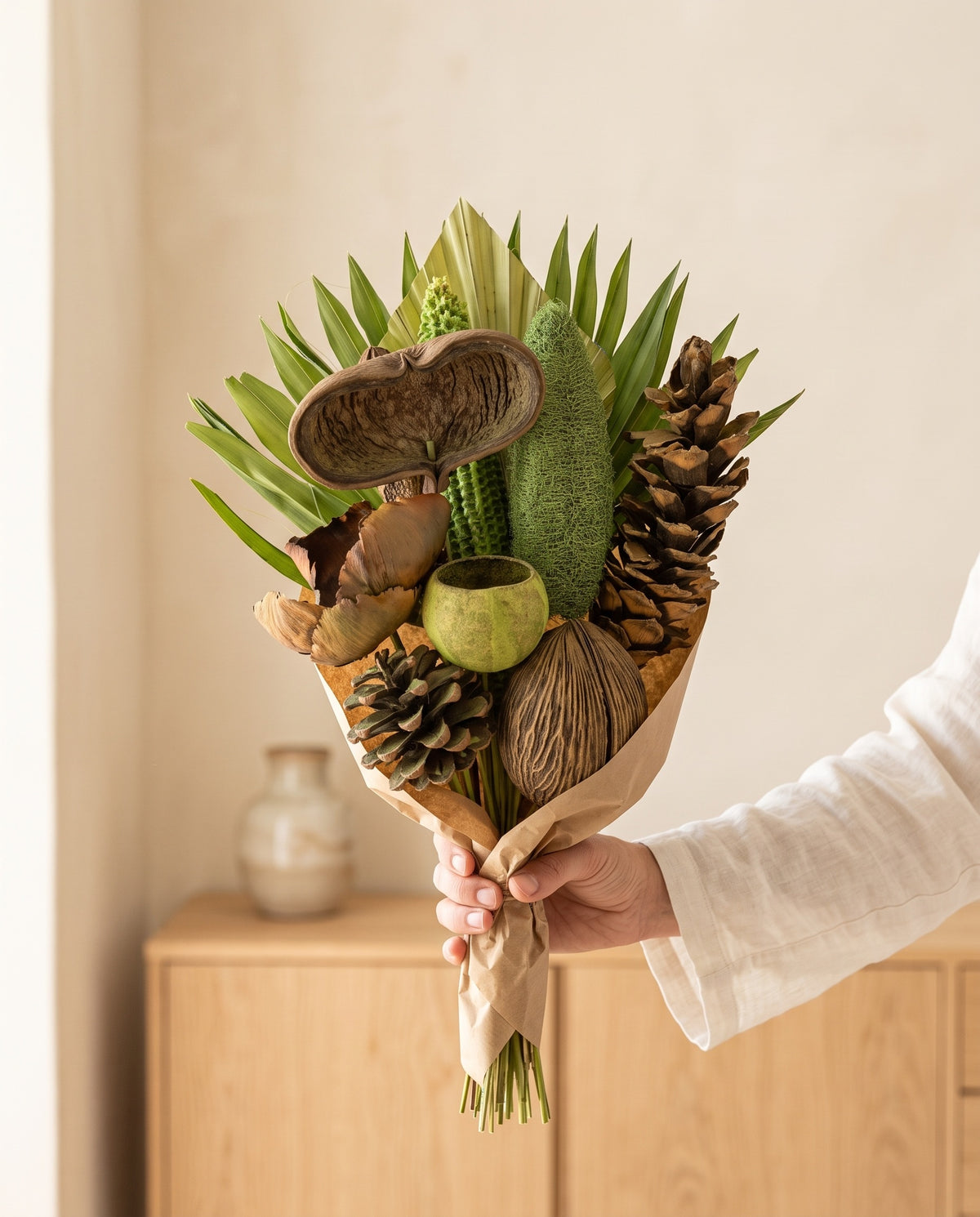 Mão a segurar o ramo de flores secas em verde-pistácio com pinhas, para ver o tamanho.