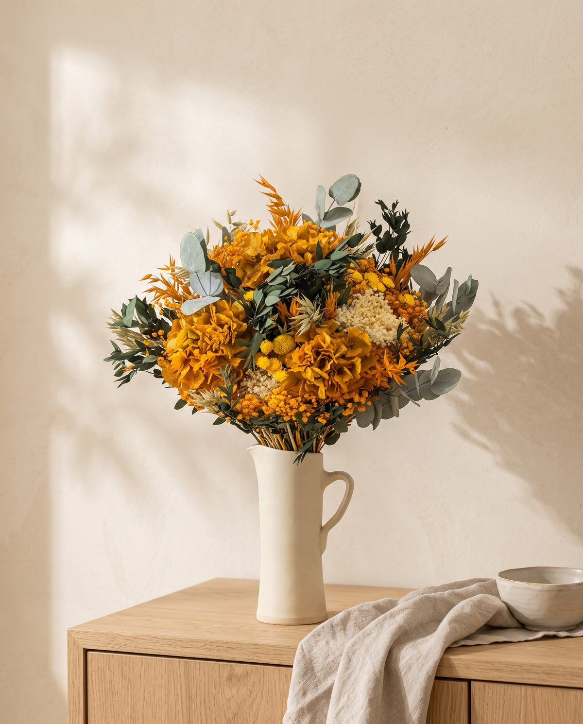 Ramo de flores secas y preservadas en amarillo y naranja en una jarra sobre un aparador, ambiente de casa. Jarra no incluida.