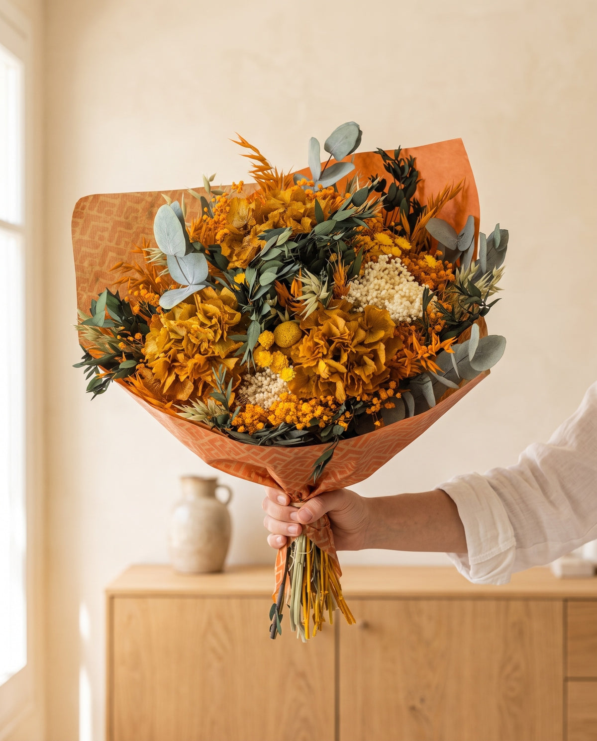 Mano sujetando el ramo de flores secas y preservadas en amarillo y naranja con su papel, como se entrega.