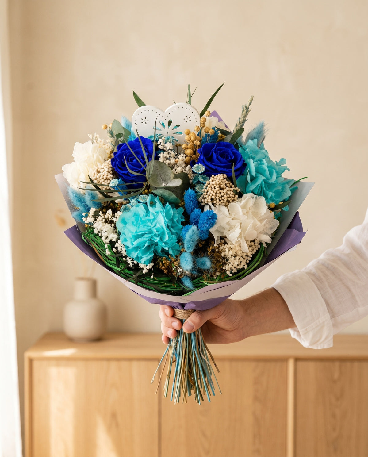 Mano sujetando el ramo de flores secas y preservadas en azul con su papel, como se entrega.