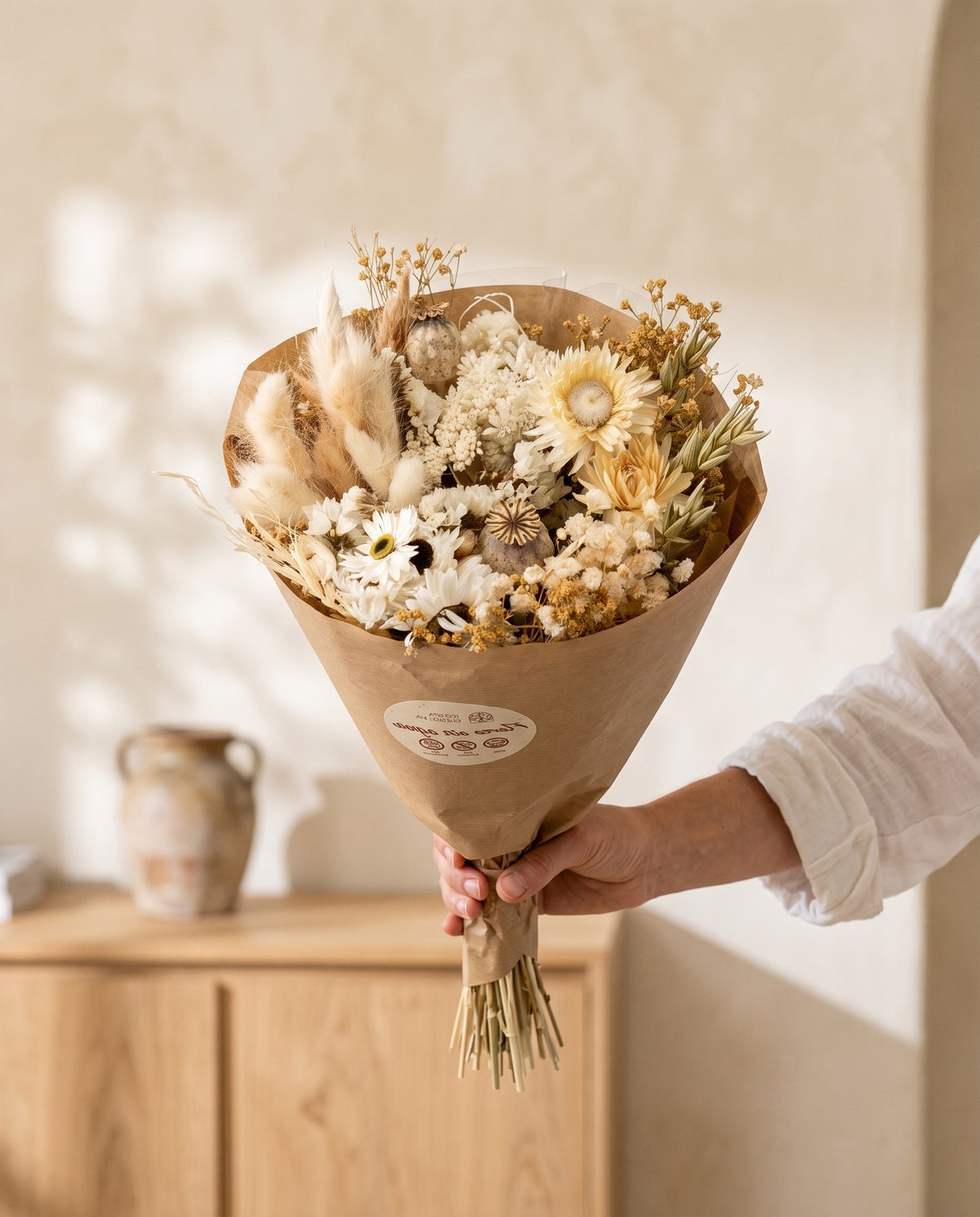 Mão a segurar o ramo de flores secas e preservadas em branco com papaver com o seu papel, tal como é entregue.