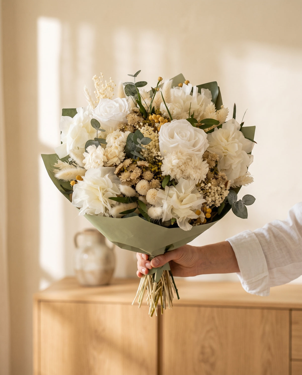 Mano sujetando el ramo de flores secas y preservadas en blanco con rosas con su papel, como se entrega.