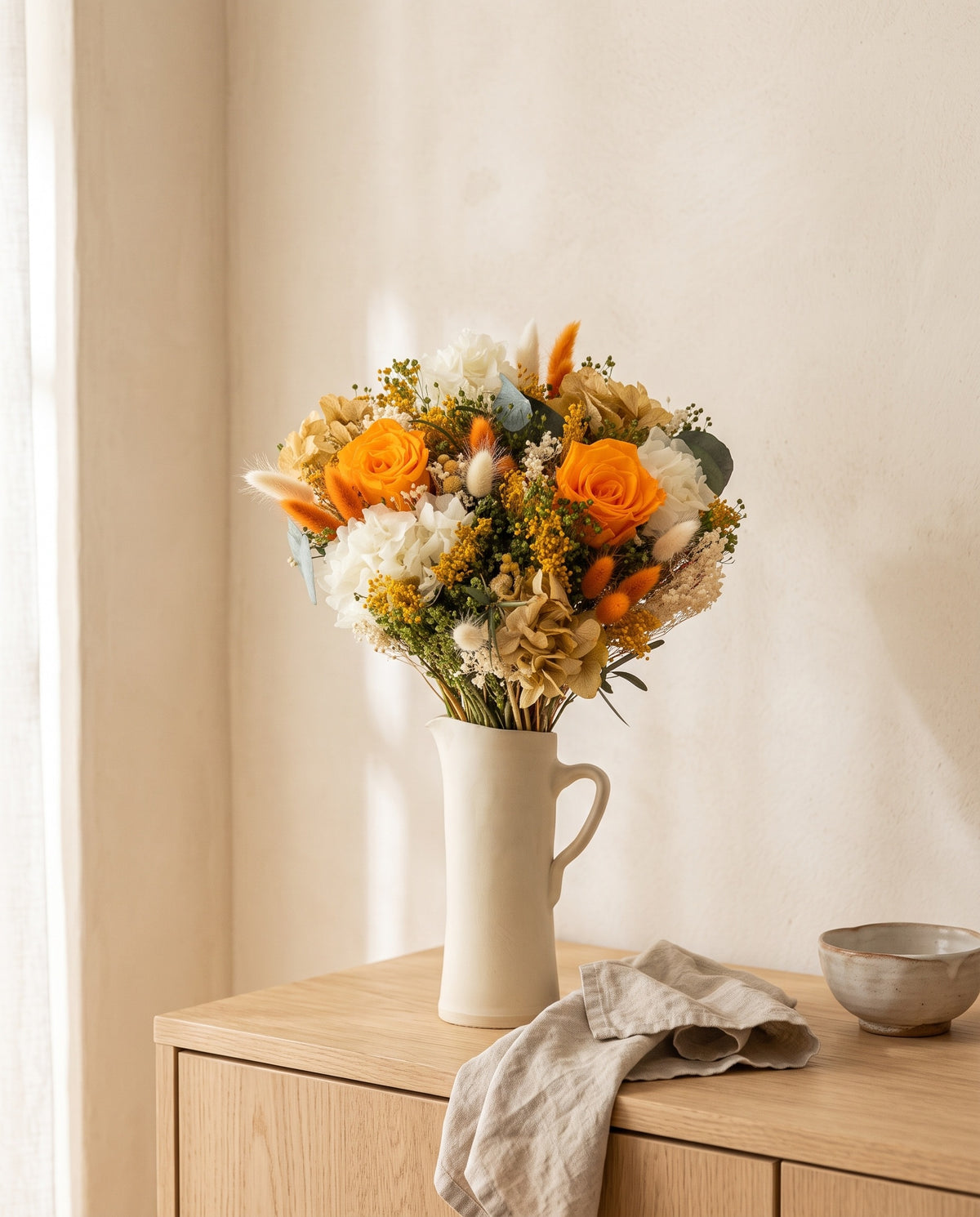 Ramo de flores secas y preservadas en blanco y naranja en una jarra sobre un aparador, ambiente de casa. Jarra no incluida.