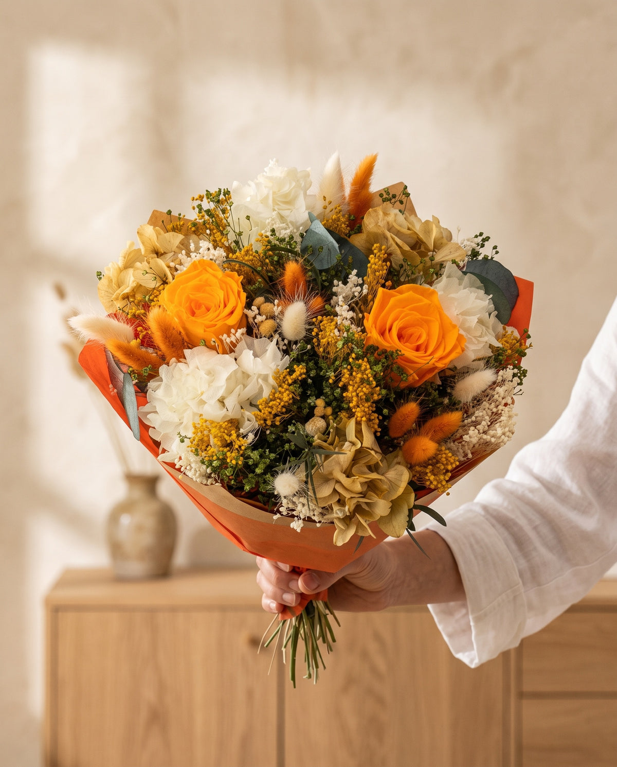 Mano sujetando el ramo de flores secas y preservadas en blanco y naranja con su papel, como se entrega.