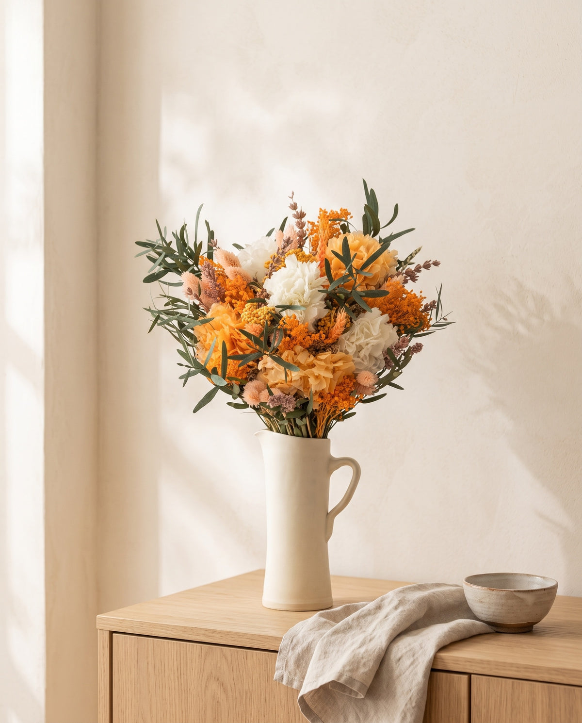 Ramo de flores secas y preservadas en blanco y naranja en una jarra sobre un aparador, ambiente de casa. Jarra no incluida.