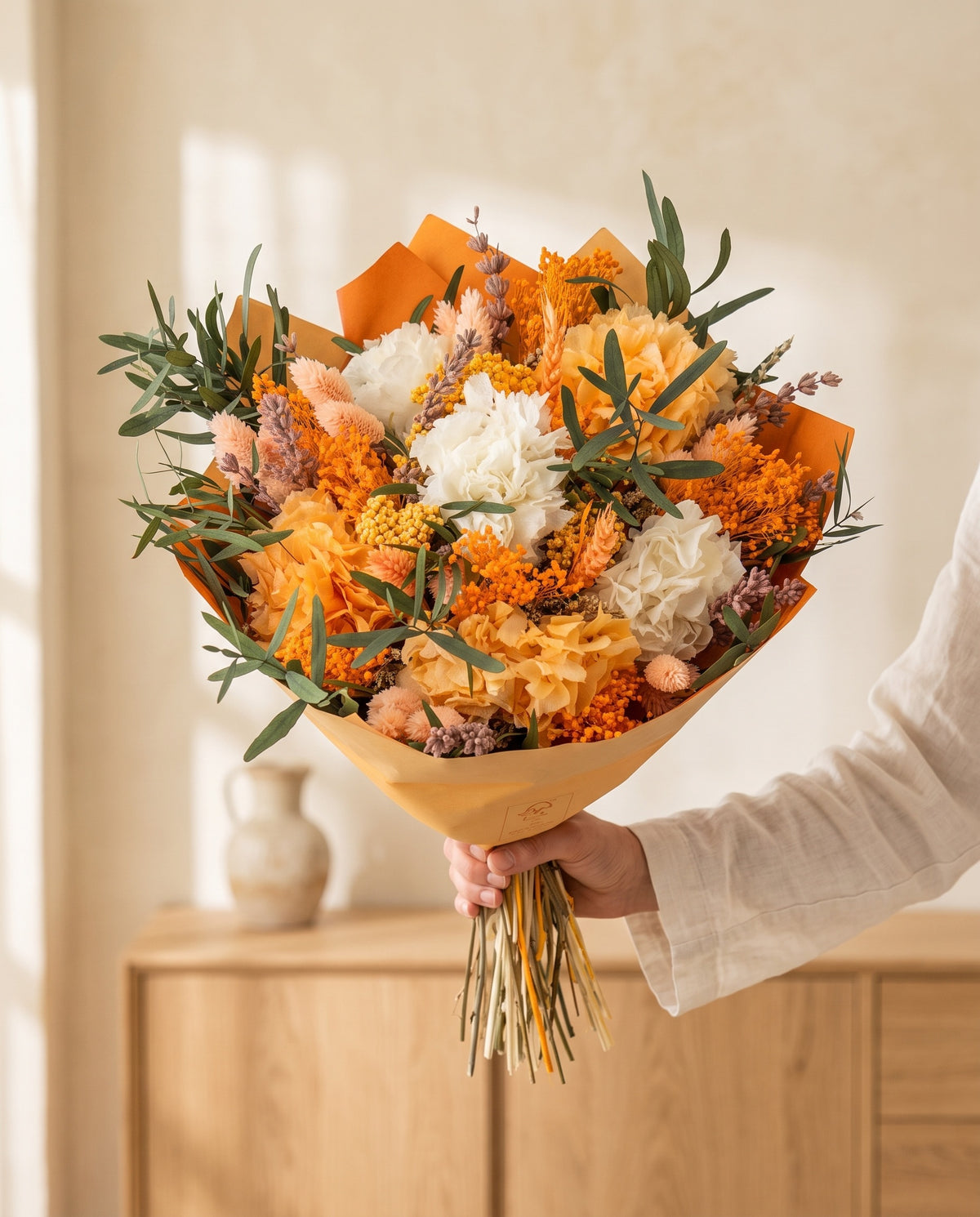 Mano sujetando el ramo de flores secas y preservadas en blanco y naranja con su papel, como se entrega.