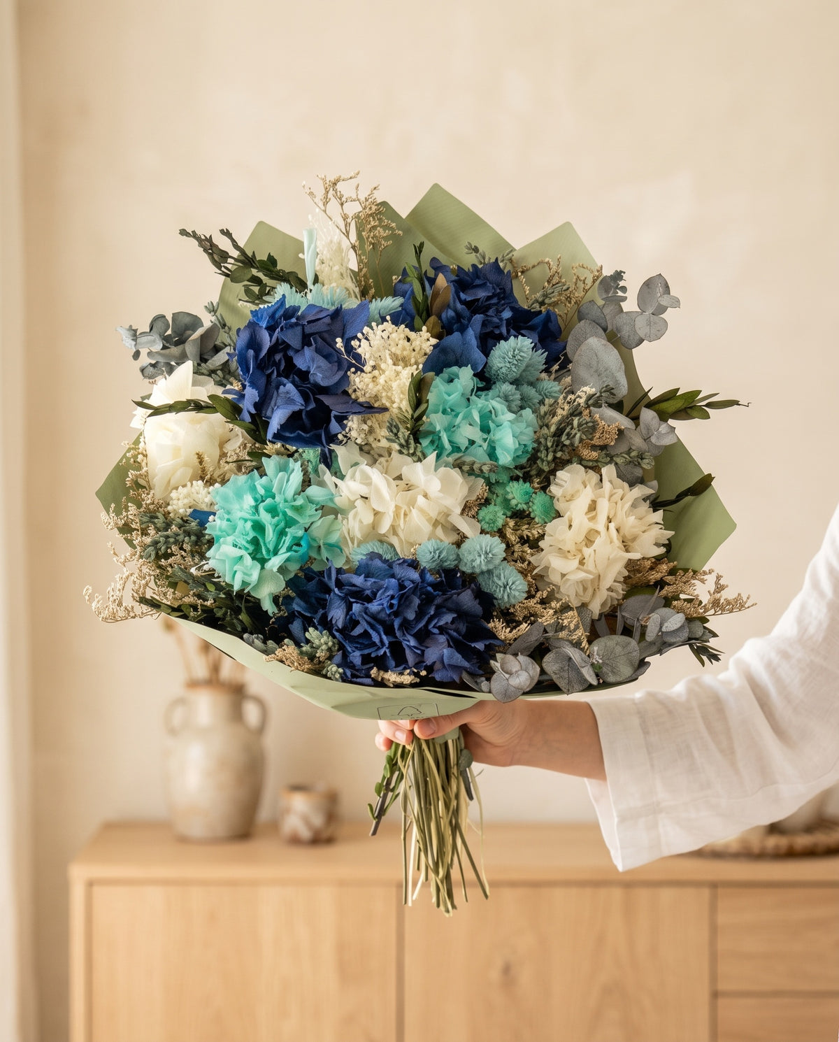 Mano sujetando el ramo de flores secas y preservadas en celeste con hortensia con su papel, como se entrega.