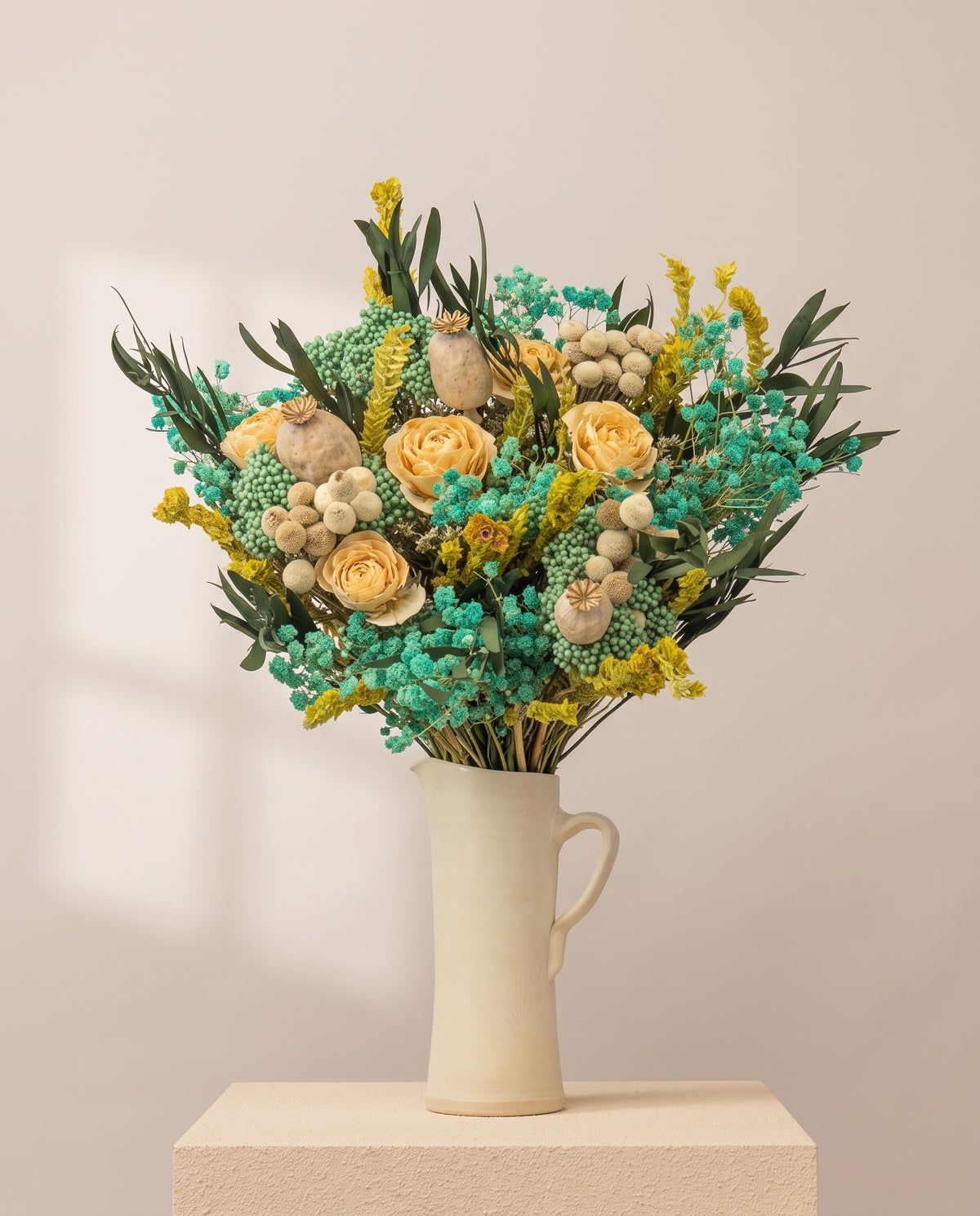 Ramo de flores secas y preservadas en celeste con rosas en una jarra de cerámica crema. Jarra no incluida.