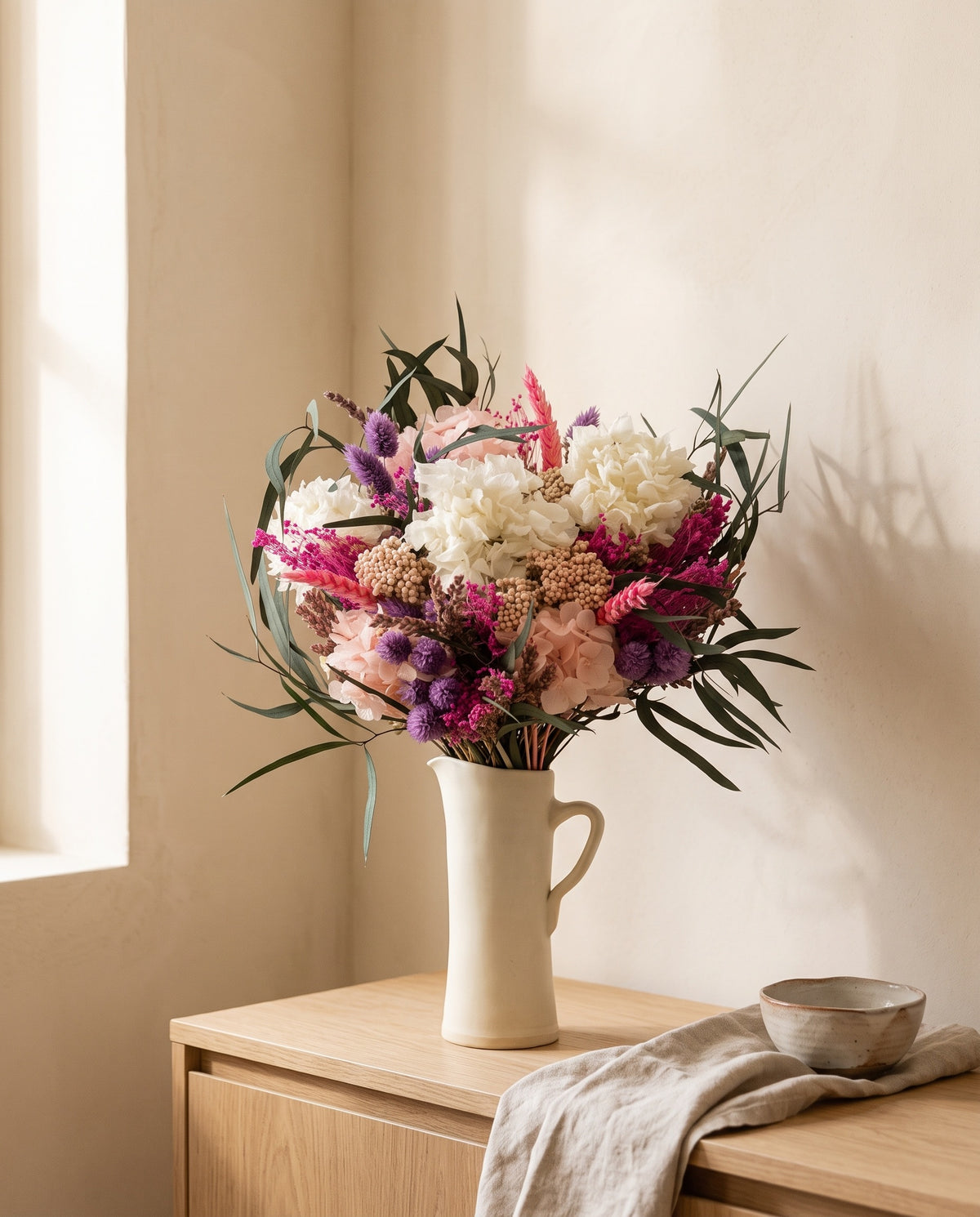 Ramo de flores secas e preservadas em fúcsia e branco numa jarra sobre um aparador, ambiente de casa. Jarra não incluída.