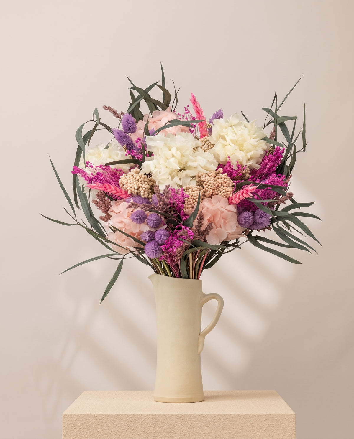 Ramo de flores secas e preservadas em fúcsia e branco numa jarra de cerâmica creme. Jarra não incluída.