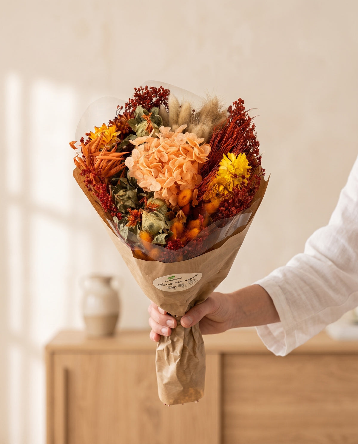 Mano sujetando el ramo de flores secas y preservadas en naranja con hortensia con su papel, como se entrega.