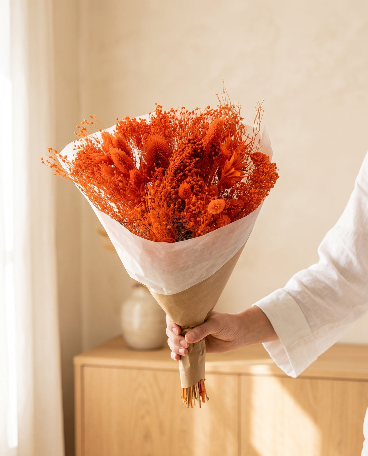 Mano sujetando el ramo de flores secas y preservadas en naranja con paniculata con su papel, como se entrega.