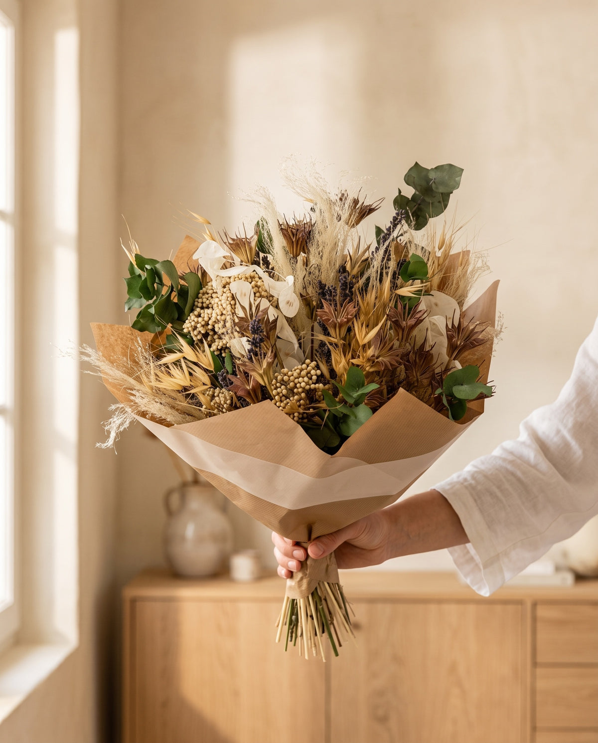 Mano sujetando el ramo de flores secas y preservadas en tonos naturales con su papel, como se entrega.