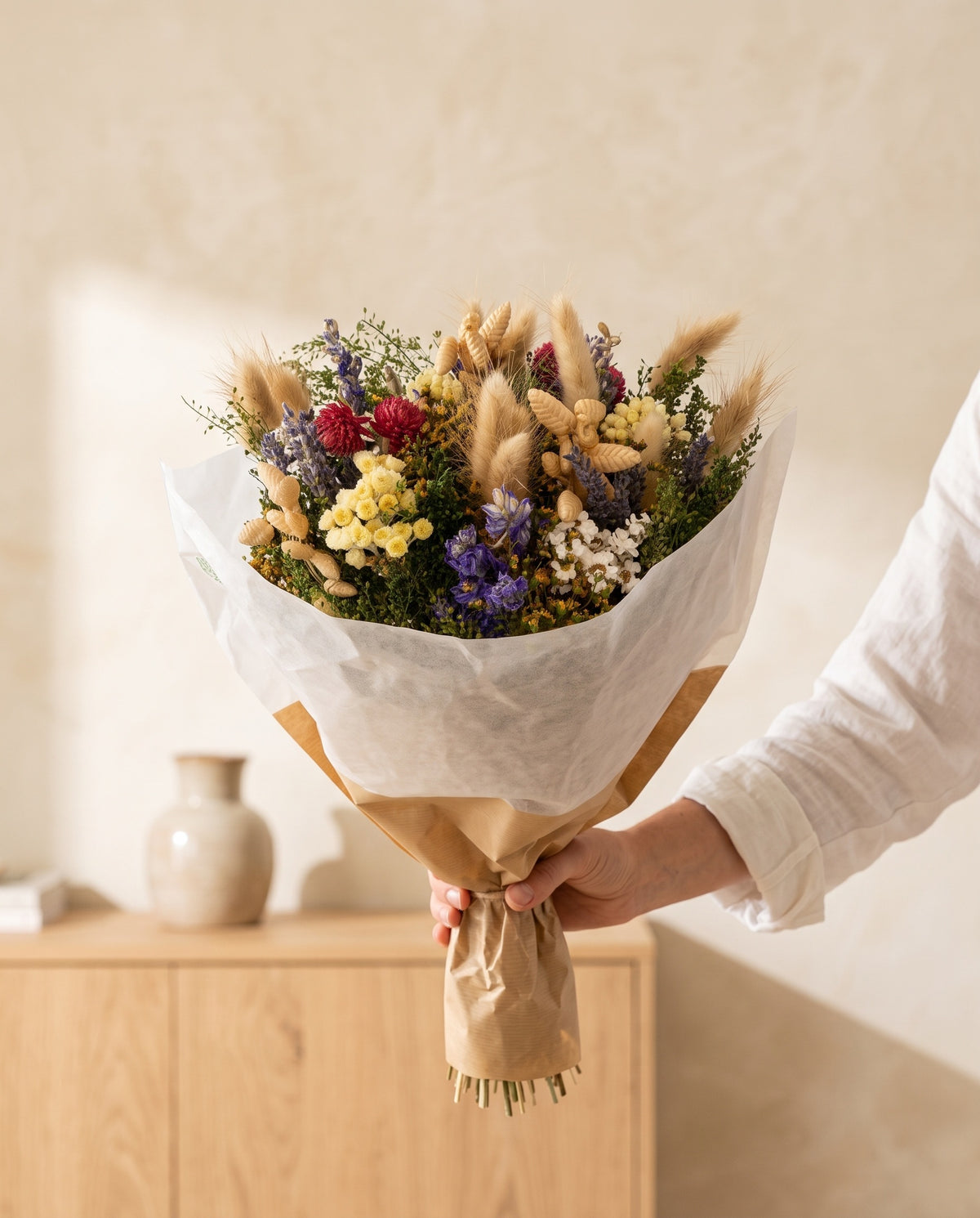 Mano sujetando el ramo de flores secas y preservadas en tonos naturales con su papel, como se entrega.