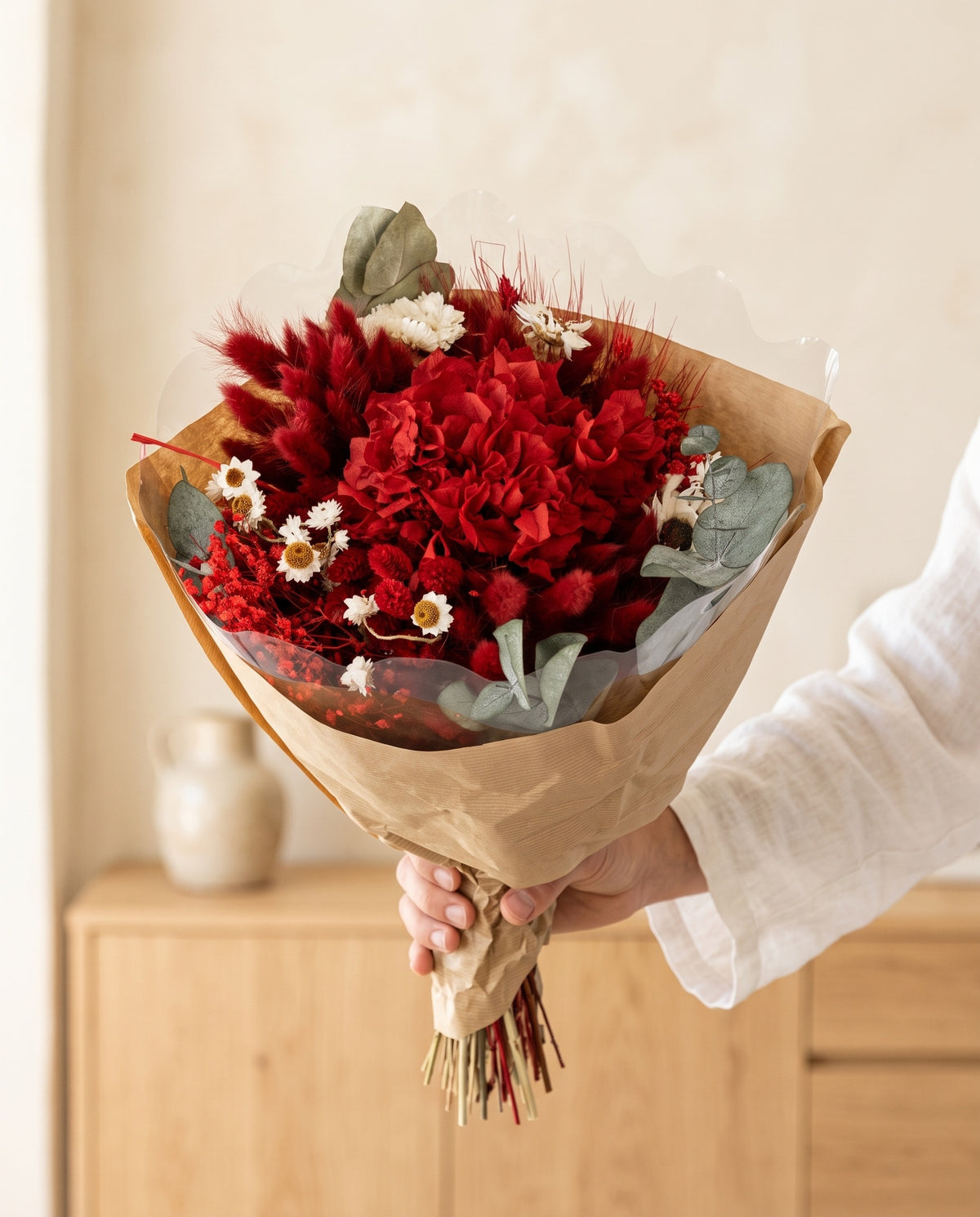 Mano sujetando el ramo de flores secas y preservadas en rojo con hortensia con su papel, como se entrega.