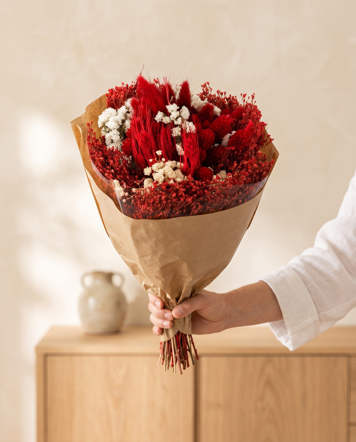 Mano sujetando el ramo de flores secas y preservadas en rojo con lagurus con su papel, como se entrega.
