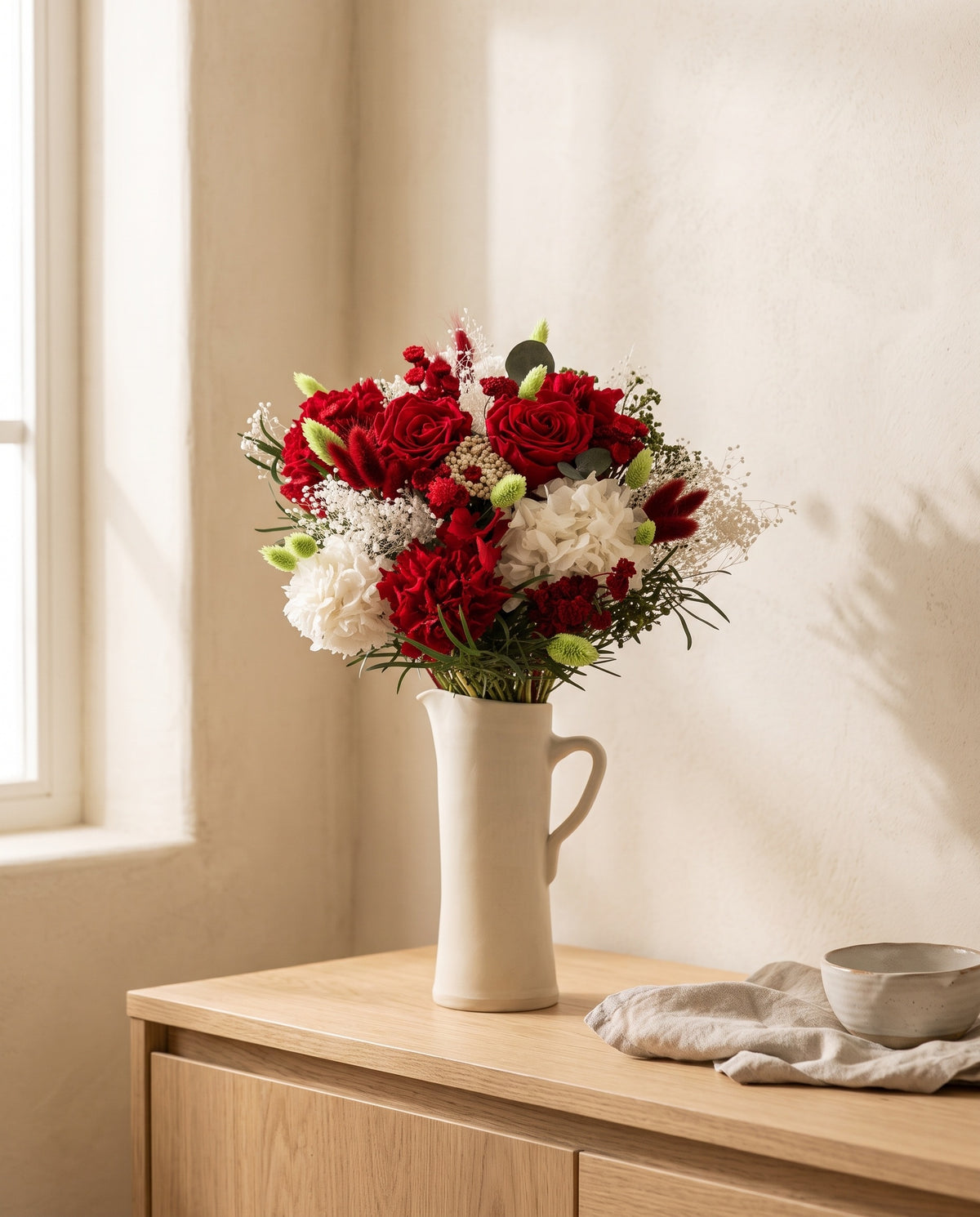 Ramo de flores secas y preservadas en rojo con rosas en una jarra sobre un aparador, ambiente de casa. Jarra no incluida.